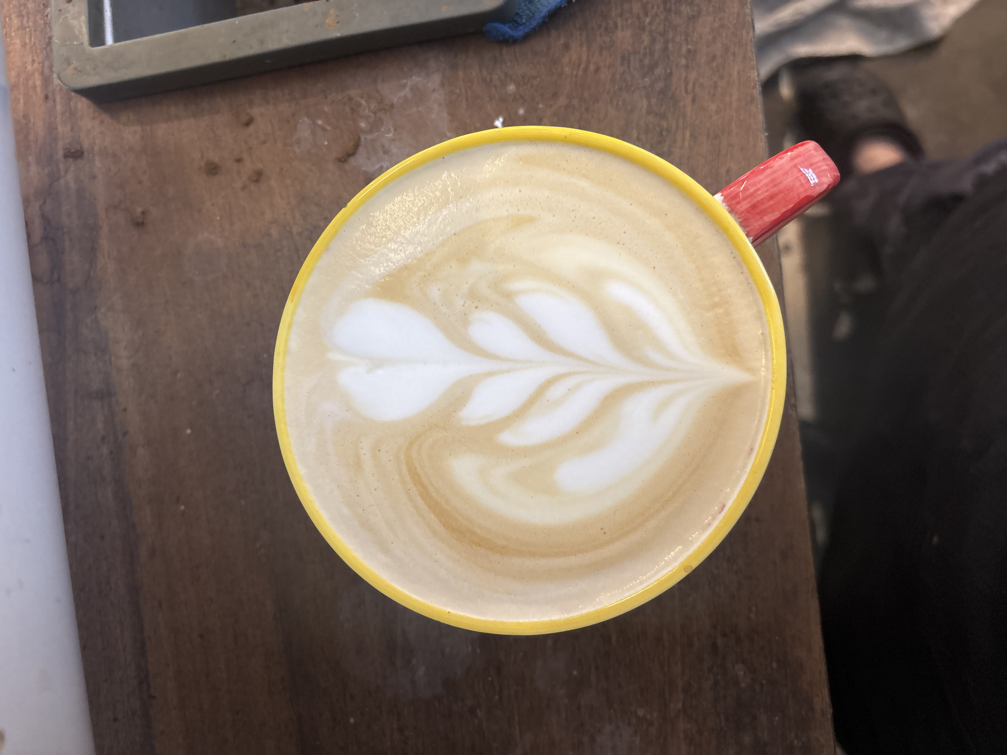 A latte with leaf art sits in a yellow mug with a red handle on a brown wooden surface. The latte art consists of white, leaf-shaped patterns against a tan background. Part of a metal object and a blue cloth appear in the upper left corner. A person wearing black pants and black shoes is partially visible on the right side of the frame. There are no indications of the location being Buenos Aires, Argentina.
