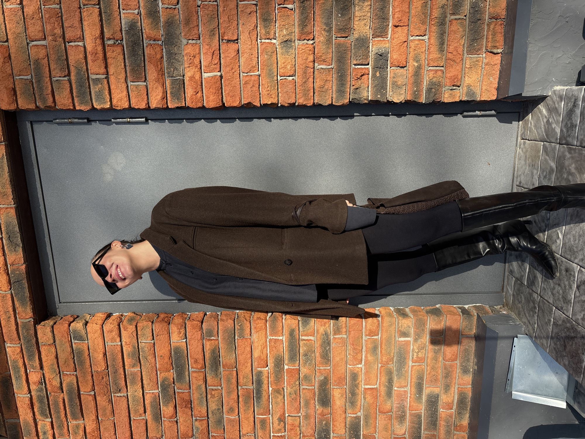 A person lies on a gray surface between brick walls. The person wears a brown coat, black clothing underneath, black sunglasses, and black boots. The location appears to be an exterior setting, possibly in Chaikovsky, Russia. The photo is taken at an angle, making it appear as if the person is lying horizontally. The environment includes a gray surface, brick walls, and tiled surfaces.