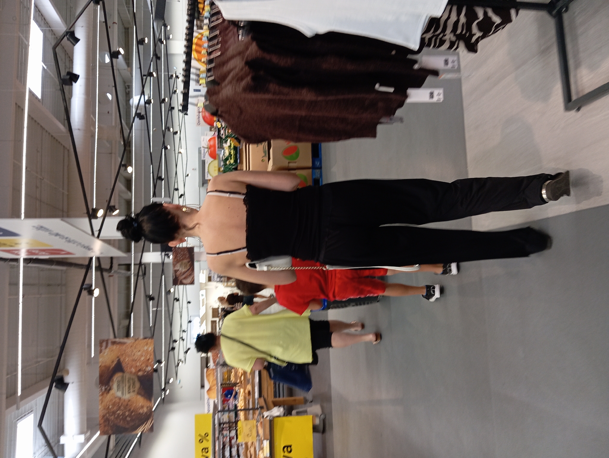 The image depicts an indoor scene, likely a retail environment such as a supermarket or department store, with a focus on clothing racks and grocery aisles. In the foreground, a woman with dark hair tied up is walking away from the camera, wearing a black tank top with lace trim and black pants. She is also carrying a white purse over her shoulder. To her left, a young boy in a red t-shirt and red shorts is walking ahead of her, pushing a shopping cart. Further to the left, a woman in a bright yellow, loose-fitting t-shirt and dark shorts is walking with her back to the camera, carrying a blue bag over her left shoulder. The background shows shelves stocked with goods, including food items and clothing. The ceiling features modern lighting fixtures and a ventilation system. The overall environment suggests a commercial setting, with a focus on shopping. The scene is set in Nitra, Slovakia, as indicated by the presence of Tesco stores, a common supermarket chain in the region.
