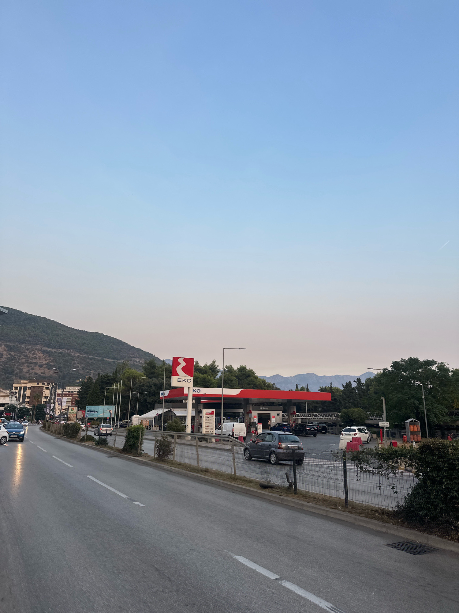 The image captures a scene in Budva, Montenegro, with a prominent EKO gas station visible on the right side. The gas station features a red canopy with EKO branding, illuminated by its signage and the lights of the pumps. Several cars are present at the station, including a grey hatchback, a white SUV, and a dark-colored vehicle. In the background, a mountain range forms a scenic backdrop to the urban environment. The foreground shows a road with traffic, including a blue car on the left and a grey car approaching the gas station. The sky is clear and a pale blue, indicating it is either early morning or late evening. The overall impression is a typical roadside scene in a coastal city.