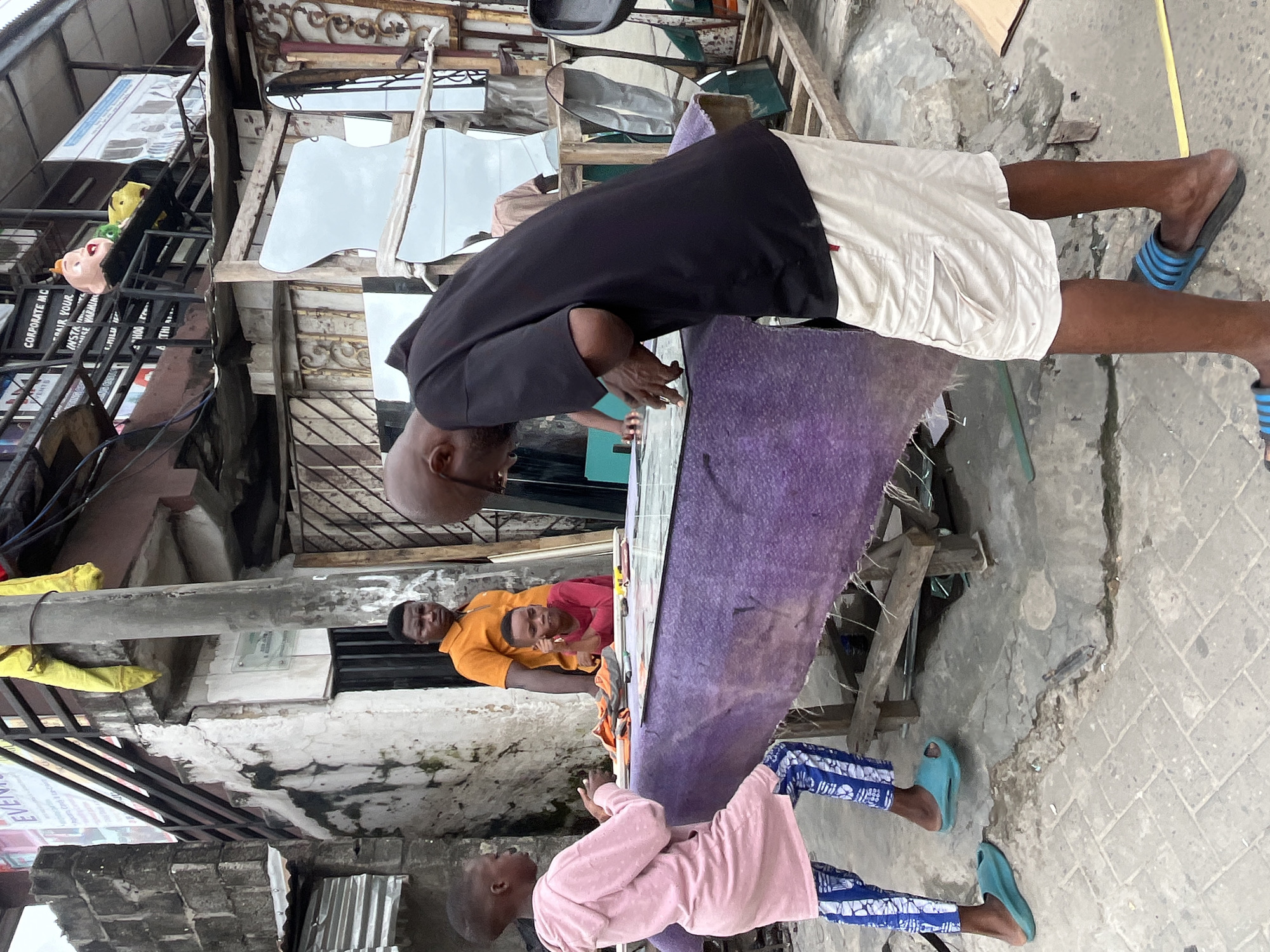 In Ado, Nigeria, a group of individuals are gathered around a table displaying a large pane of glass. A man in a black t-shirt and white shorts is actively working on the glass, seemingly measuring or cutting it, with his hands positioned on the surface. Beside him, a young person in a pink hooded garment and patterned blue and white pants stands with their hands clasped, observing the process. Behind them, a man in an orange shirt looks on, while a younger individual in a red shirt sits at the table, also attentive to the task. The environment appears to be an open-air market or workshop, with various mirrors and building materials visible in the background. The ground is paved with interlocking bricks and shows some signs of wear and tear.