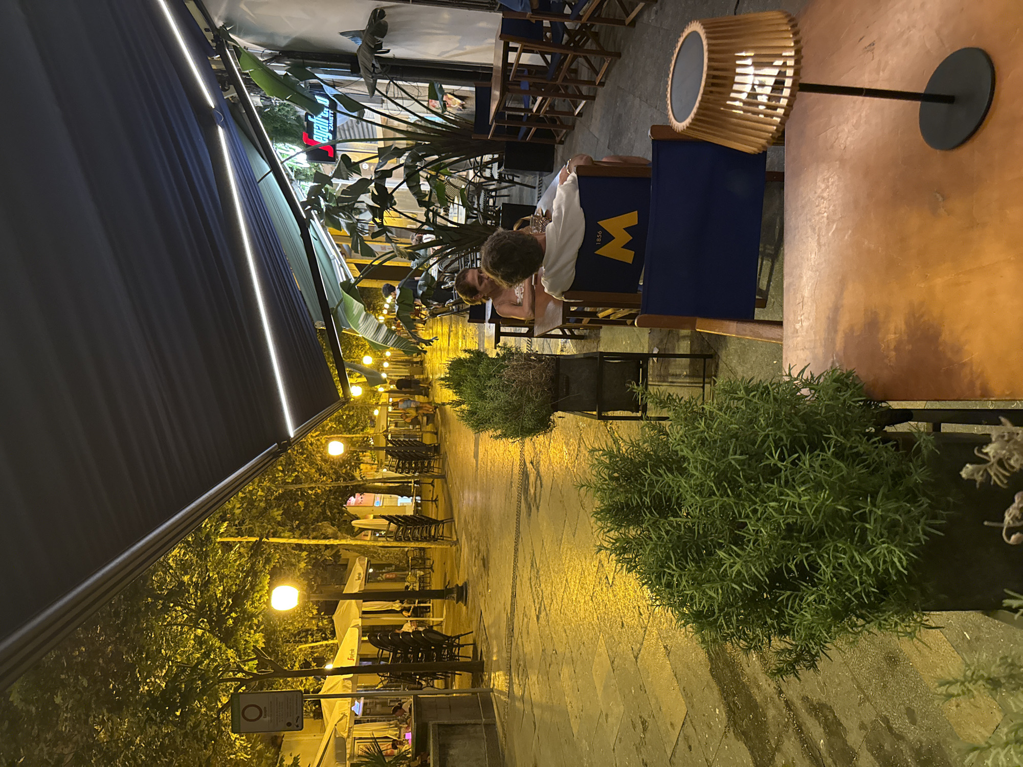 This image depicts an outdoor dining area at night in Blanes, Spain. The scene is illuminated by streetlights and the lights from an awning. The wet cobblestone street reflects the yellow light, suggesting it may have rained recently. Several tables and chairs are set up outside a restaurant, with some tables occupied by patrons. In the foreground, a large potted plant with green foliage is visible. Further back, a couple is seated at a table, and other patrons can be seen at distant tables. Chairs are stacked on some of the tables, indicating that the restaurant may be closing or has a specific setup for seating. A sign with red lettering is visible on the awning.