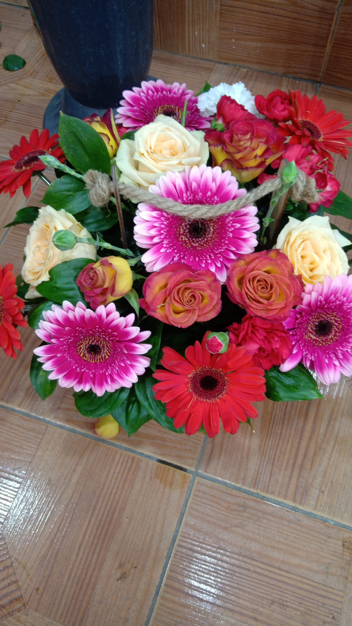 A close-up shot from above shows a vibrant bouquet of flowers arranged in a container. The flowers include pink and red gerbera daisies, peach and orange roses, a single white carnation and smaller buds. The stems are wrapped in a rough, twine-like material. Green leaves fill the spaces between the flowers. The bouquet sits on a surface of light-brown tile. The location is Yerevan, Armenia.