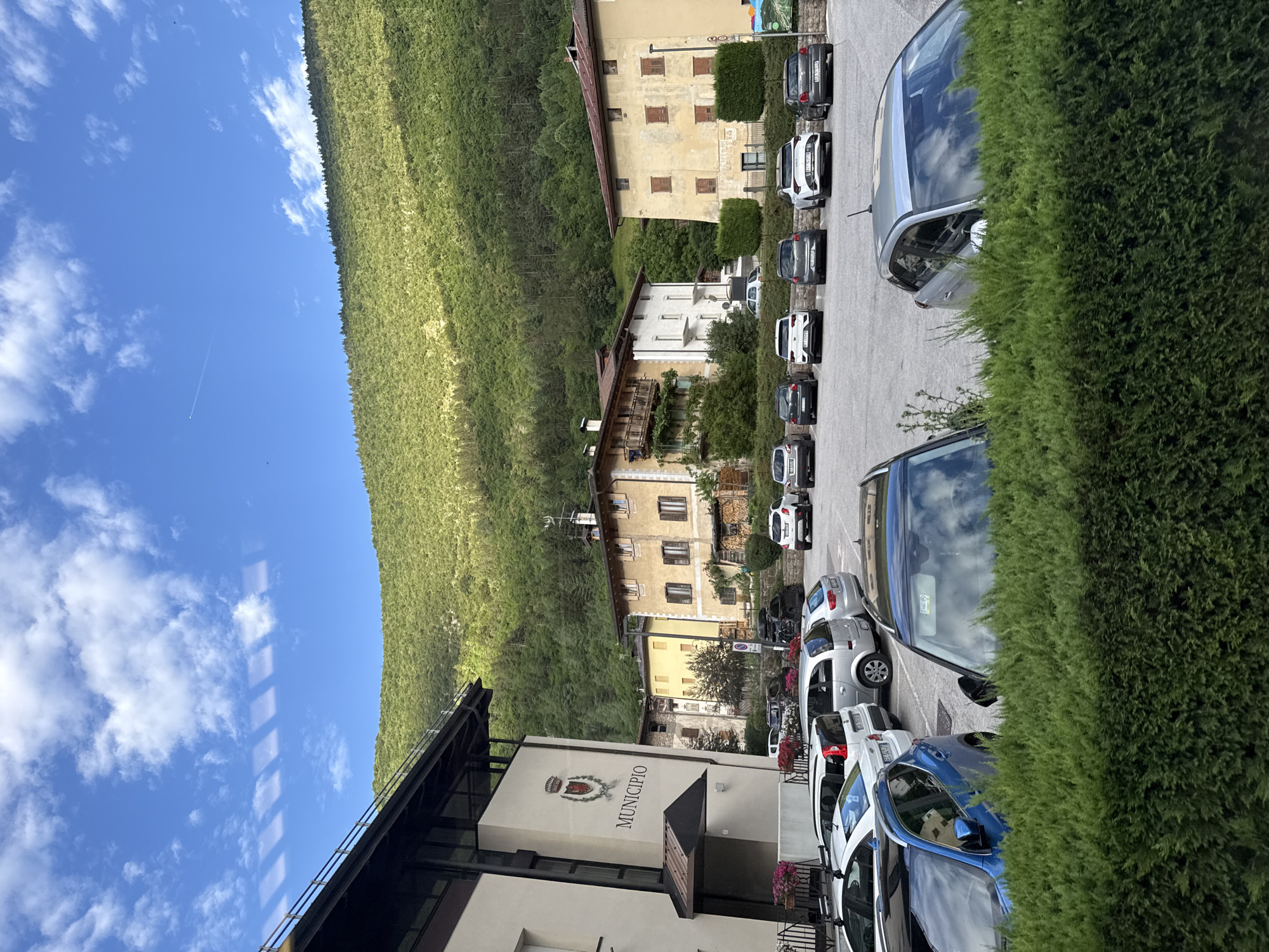 This media file shows a street scene in Spormaggiore, Italy. The image is oriented vertically, with the sky and a green, forested hillside occupying the upper portion, and a paved area with parked cars and buildings in the lower portion. Several cars are parked in a line along a paved surface. The buildings appear to be a mix of residential and commercial, with one building clearly marked "MUNICIPIO" with a crest above it, indicating it is the town hall. Some of the buildings have balconies and wooden shutters. The sky is a clear blue with scattered white clouds. The sun appears to be shining, suggesting it is daytime. The presence of a jet contrail in the sky indicates the time is likely midday or afternoon. The overall scene depicts a quiet, possibly rural European town on a clear day. There is no clear activity or event taking place, beyond the cars being parked.