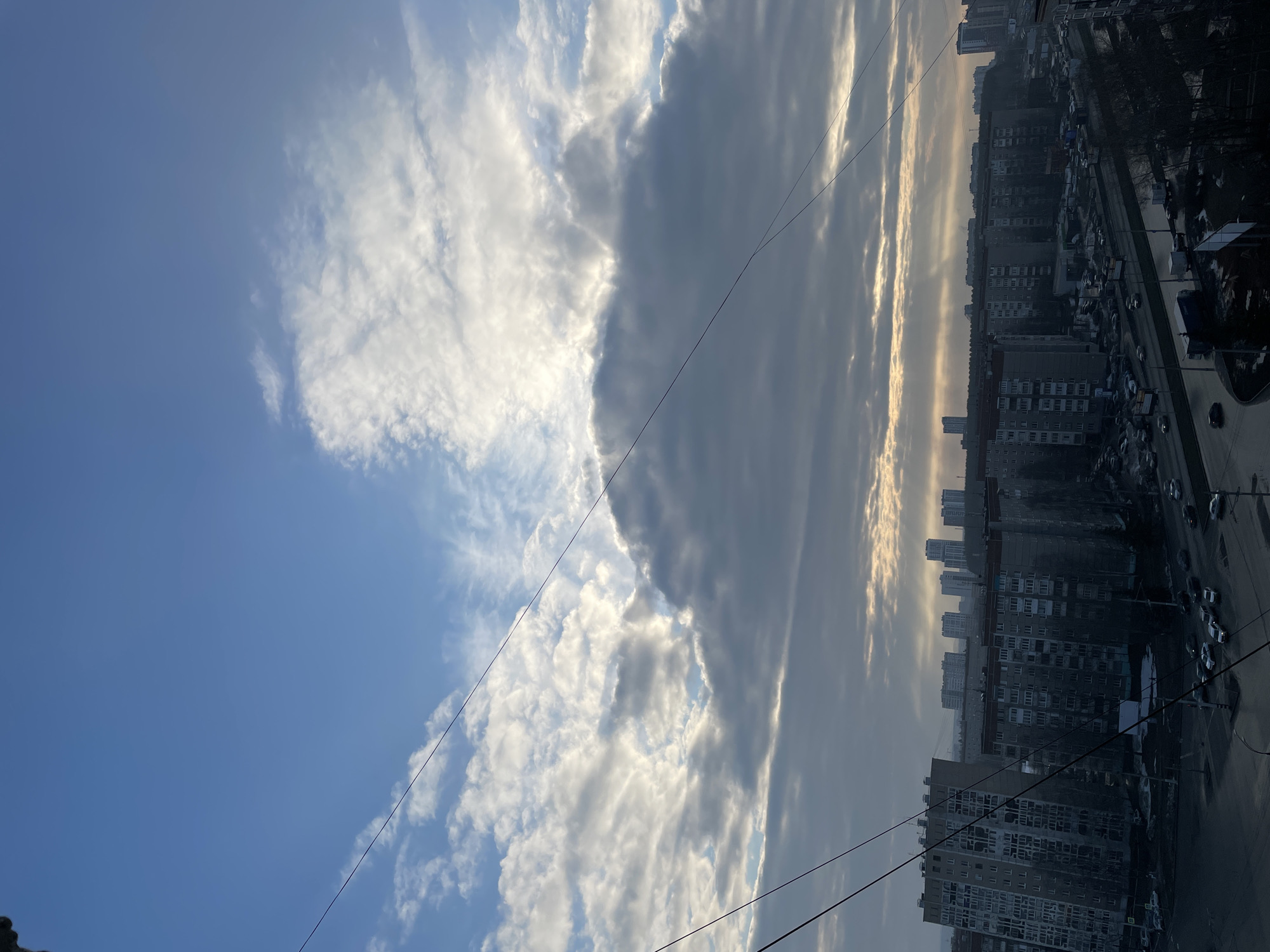 The image shows a view of Izhevsk, Russia, from a high vantage point, likely a balcony or window. The sky occupies the left portion of the frame, showcasing blue skies with scattered white clouds transitioning to darker, overcast clouds towards the right. Below the clouds is a cityscape dominated by densely packed apartment buildings. The buildings appear to be of varying heights, with a monotonous gray facade. Streets are visible, with parked cars and sparse traffic. The overall atmosphere is one of urban density under a partly cloudy sky. Power lines run diagonally across the frame.