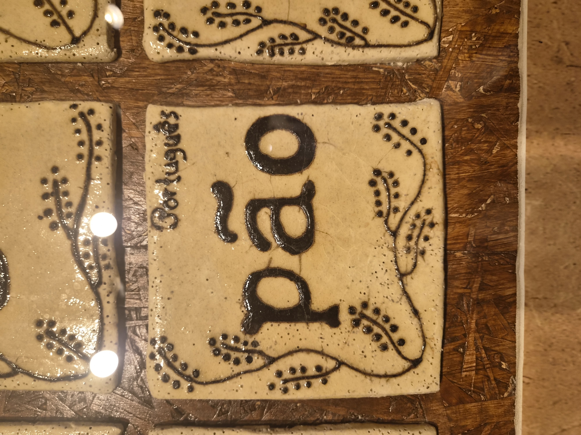 The image shows three ceramic tiles on a wooden surface. The central tile features the word "pão" in large, dark letters, with a tilde above the 'a'. Above "pão" and on the tile to its right, the word "Português" is written in smaller letters. All three tiles are decorated with dark, line-drawn botanical motifs, resembling branches with small berries or dots. The tiles are arranged in a horizontal row, with the central tile being the most prominent. The background appears to be made of pressed wood chips. The ceramic tiles have a light, speckled glaze. Two bright white circular reflections are visible on the surface of the tiles, likely from a light source. The scene is an exterior shot of a display of artisanal tiles. The overall impression is one of rustic craftsmanship and cultural representation, possibly in a shop or market stall. The context of Seia, Portugal, suggests a connection to Portuguese culture and artisanal traditions.