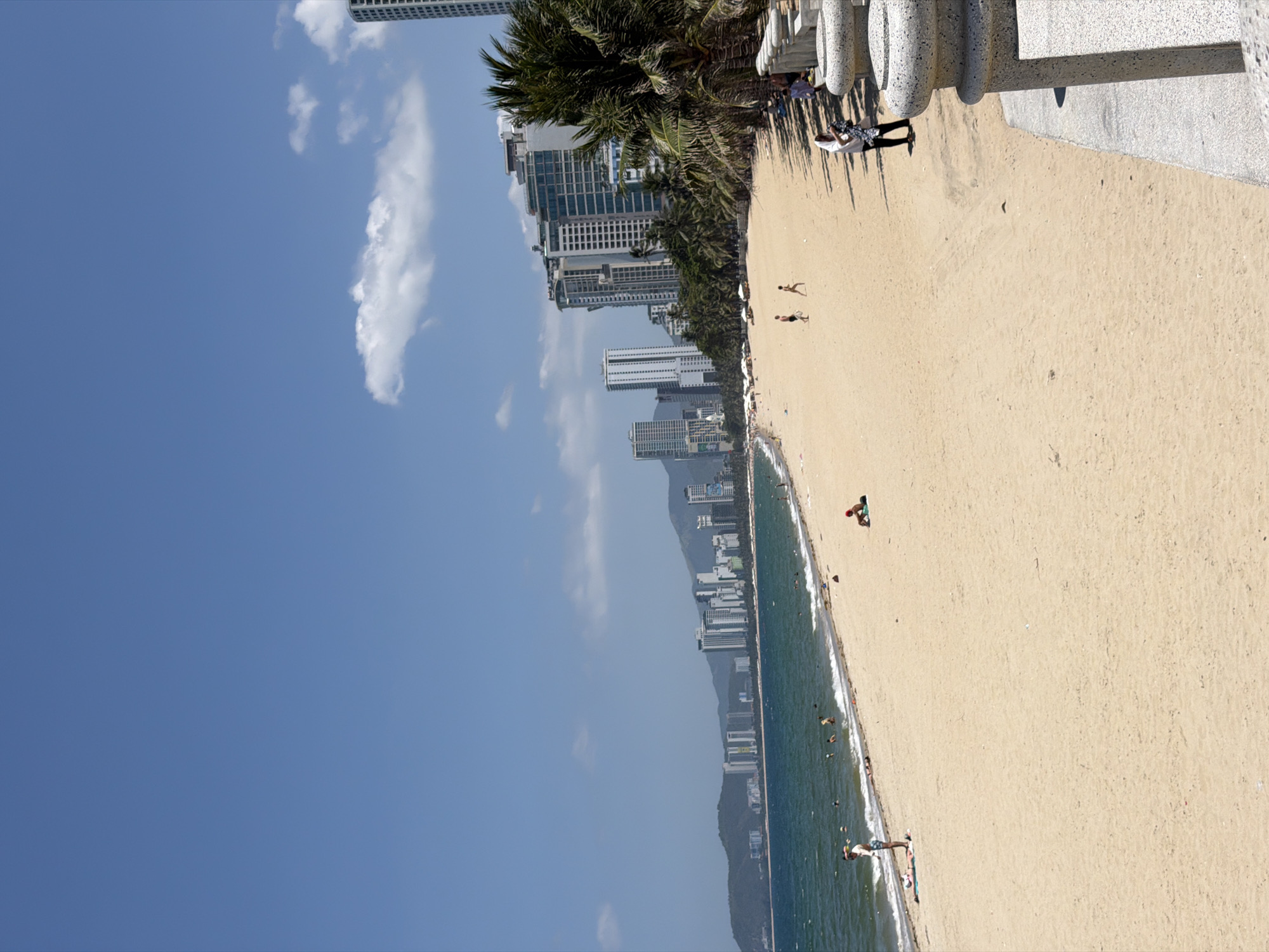 A wide-angle, eye-level photograph captures a sunny daytime scene of a beach along the coast of Nha Trang Ward, Vietnam. The image is horizontally divided into approximately one-third sky, one-third ocean/cityline, and one-third beach.
The upper portion features a bright blue sky with a few sparse white cumulus clouds. Below, the turquoise-blue ocean extends towards a distant horizon, showing gentle waves breaking near the sandy shore. Approximately ten individuals are visible swimming or wading in the ocean.
Along the coastline, a dense city skyline is present, composed of numerous modern high-rise buildings with light-colored facades and extensive windows. These structures are backed by dark green mountains in the far distance on the left. A line of tall palm trees runs parallel to the beach, separating the sand from the urban development on the right side of the frame.
The lower portion of the image is dominated by a wide expanse of light golden-beige sand. On the left side of the beach, one adult male in a light shirt and shorts stands near the water's edge, while another individual lies prone on a blue towel. In the mid-ground, two figures, possibly a child and an adult, walk on the sand. Further to the right, one person reclines on a red towel.
On the far right, a concrete promenade with a grey stone railing featuring rounded posts is partially visible. One individual, dressed in a white long-sleeved top and dark pants, stands near the railing, holding a dark garment. Shadows from the palm trees and railing extend onto the sand and promenade, indicating strong overhead sunlight. Several indistinct figures are seated or standing in the shade under the palm trees further right.