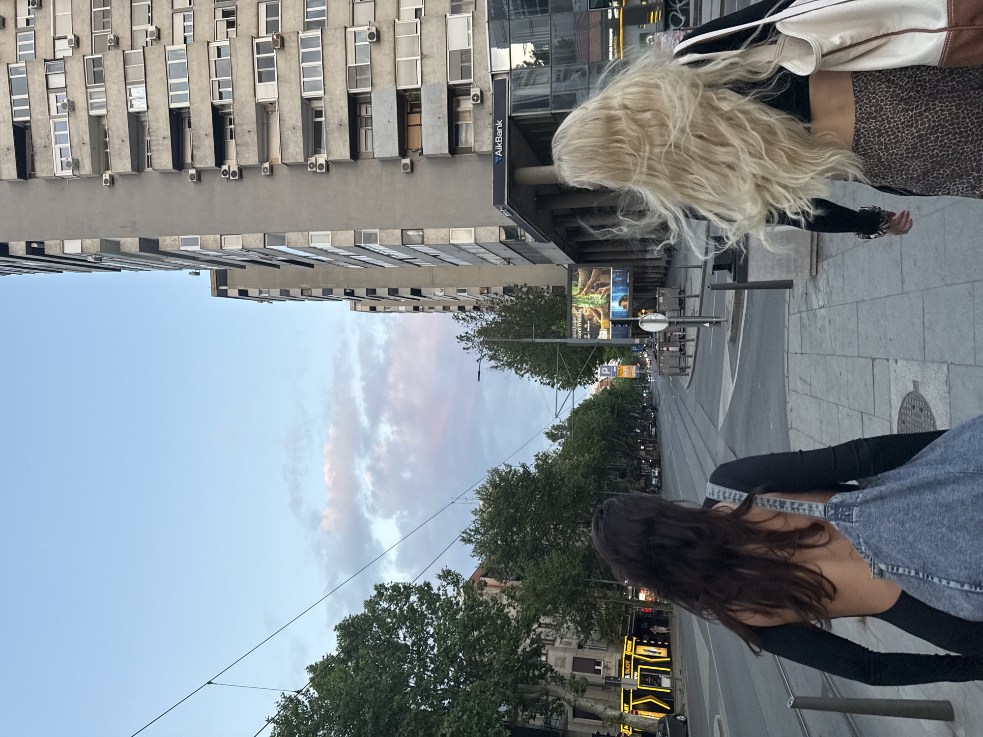 A medium shot shows two women walking on a city sidewalk, in Palilula Urban Municipality, Serbia. The woman on the left has long dark hair and is wearing a dark long-sleeved shirt and a denim jumper. The woman on the right has long blonde hair and is wearing a dark long-sleeved shirt and a patterned skirt. A white bag is slung over her shoulder.
A tall, gray building with many windows and air conditioning units is on the left side of the image. A sign for "AIK Bank" is visible on the side of the building. A street with trees lining it stretches into the distance. The sky is a mix of blue and pink. Street lamps and traffic signals are visible along the street. Several businesses with illuminated signs are on the right side of the street in the background. The sidewalk is made of gray concrete slabs. Tram lines run along the edge of the sidewalk.