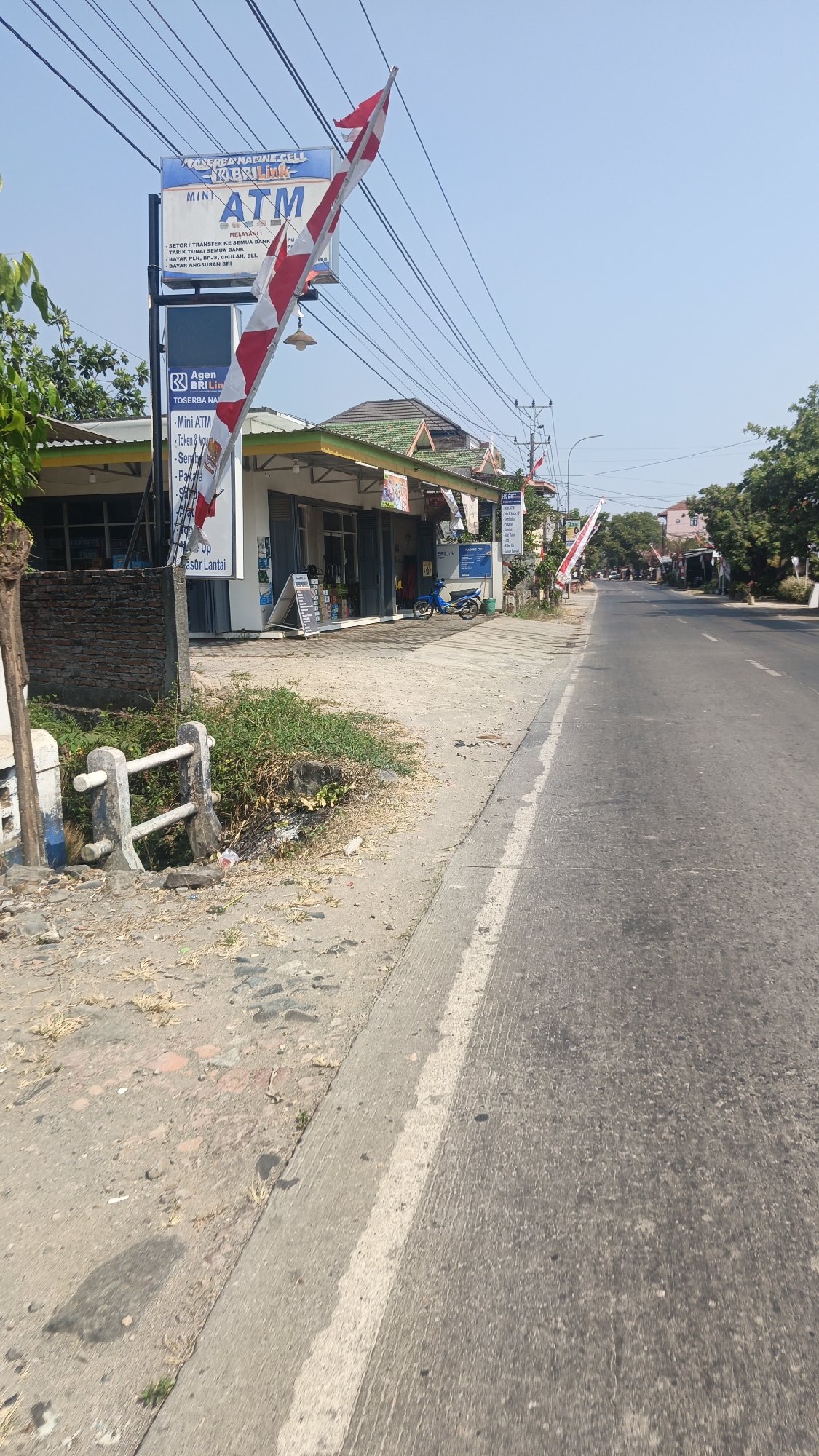 A street-level, eye-level, outdoor, medium shot captures a row of shops and a road in Jekulo, Indonesia, under a clear, pale blue sky. A prominent sign for "TOSERBA NANDINI CELL MINI ATM" stands tall, adorned with a red and white Indonesian flag. The shops, with their green tiled roofs and white facades, are open to the street. A blue motorcycle is parked in front of one of the shops. To the left of the shops, a small concrete barrier with white railings lines the edge of the sidewalk. The road, paved with asphalt, runs diagonally across the right side of the frame, with a white line marking the edge. A few trees are visible in the distance. The ground is a mix of dirt, gravel, and some sparse vegetation. The scene appears to be during daylight hours, with the sun casting subtle shadows.