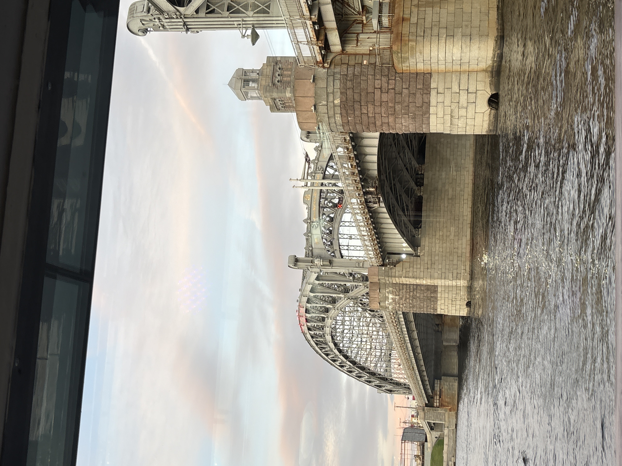 This image shows a view of the Bolsheokhtinsky Bridge in Saint Petersburg, Russia, from the Neva River. The bridge is a large, ornate metal arch bridge with stone piers. The sky is overcast with some pink and orange hues from the setting sun. The river is choppy with reflections of the bridge. The photo is taken from a boat.