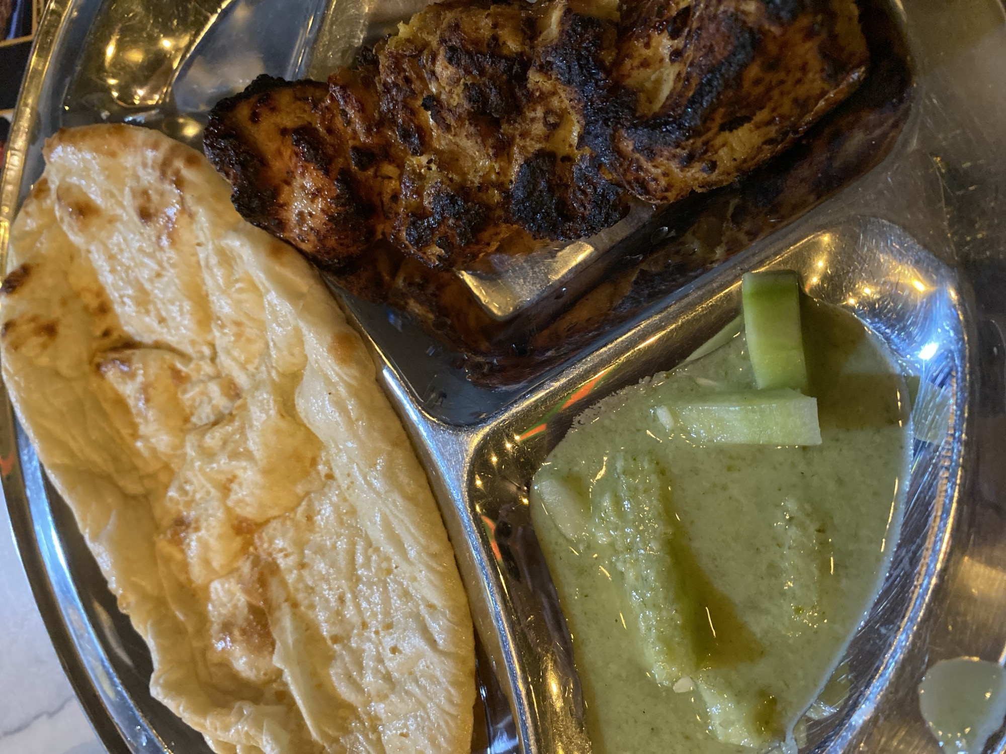 A close-up shot captures a meal served in a shiny, metallic, compartmentalized tray. The tray, possibly stainless steel, reflects ambient light and hints at an indoor setting.
In the top right section of the tray, there is a piece of grilled meat, likely chicken, characterized by its dark brown, charred exterior and visible grill marks. Its texture appears succulent and flavorful.
To the left, a golden-brown flatbread, resembling naan or paratha, occupies another section. It is soft, slightly puffed, and has a subtly uneven, browned surface, indicating a fresh bake.
The bottom right compartment holds a vibrant, light green sauce or chutney. The sauce has a somewhat smooth consistency, dotted with visible pieces of a green vegetable, which appear to be cucumber chunks, along with smaller white flecks that could be seeds or ground spices. The surface of the sauce shows slight variations in texture and some oil separation.
The meal's presentation is typical of South Asian cuisine, commonly found in regions like Dhaka, Bangladesh. The image focuses solely on the food, with no individuals present. There is no visible text. The lighting suggests an indoor environment, but the time of day is not discernible.