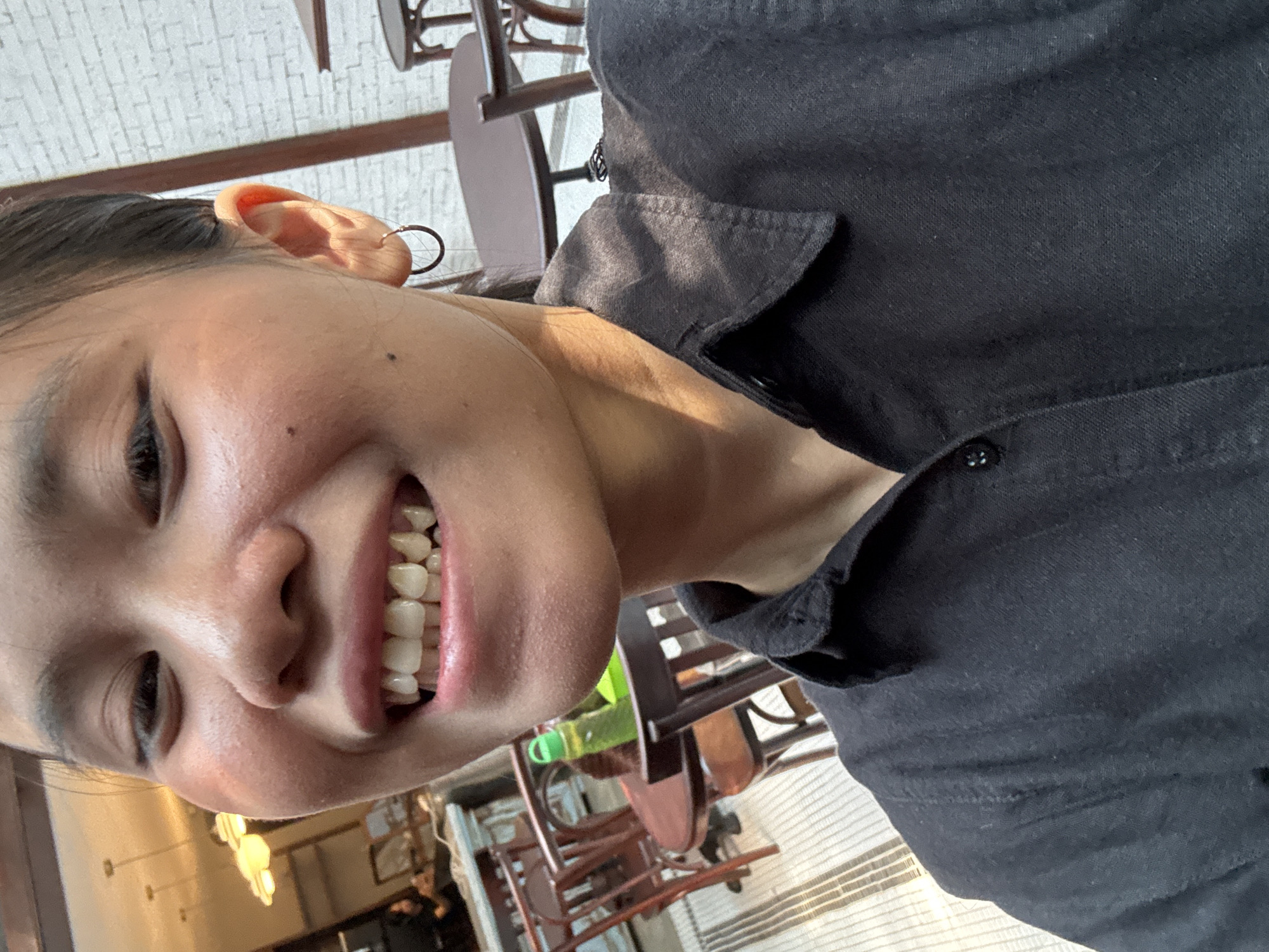 A close-up selfie of a young woman is shown, framing her face from the nose down, her dark hair pulled back. She has dark eyes, a mole on her left cheek, and is smiling, revealing her teeth. A simple hoop earring is visible on her right ear. She wears a black collared shirt. The background suggests an outdoor cafe setting in South Jakarta, Indonesia, with a brick wall and outdoor seating visible.