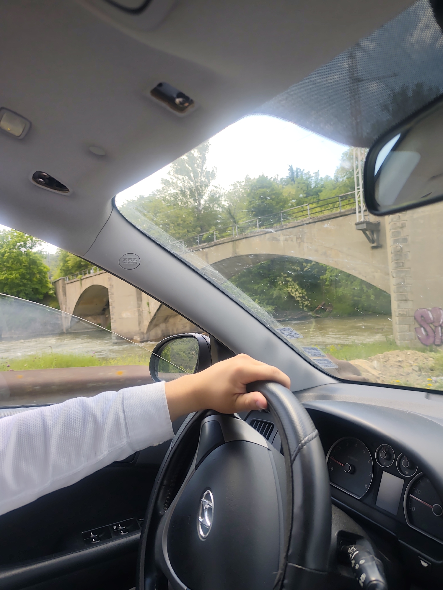 From the interior of a car, a driver grips the steering wheel with their right hand. The driver wears a white, long-sleeved shirt. Through the windshield, a two-arched stone bridge spans a flowing river. Lush greenery surrounds the bridge. Graffiti marks the side of one of the bridge's support structures. The car appears to be located in Jeleznitsa, Bulgaria, given the recognizable architecture of the bridge. The dashboard and interior controls of the car are visible, along with the rearview mirror.