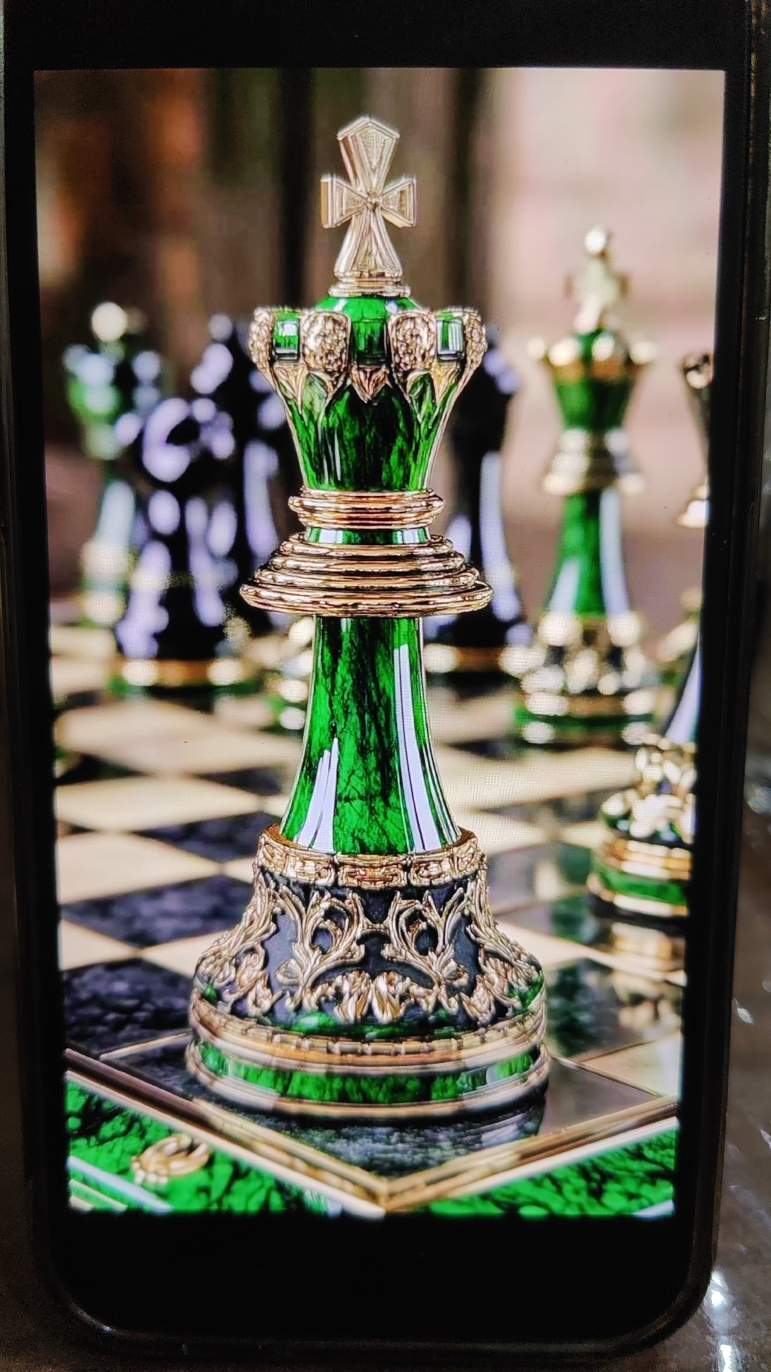 A detailed, close-up photograph showcases a striking green king chess piece, intricately designed with gold accents. The king stands on a checkered board, its base adorned with ornate gold filigree against a dark backdrop. In the blurred background, other chess pieces, including more kings and possibly queens, are visible, some also in green and gold, and others in black. The lighting highlights the rich, marbled texture of the green material. The scene is captured within a phone screen, indicated by the black border surrounding the image. There is no discernible location within the city of Sochi, Russia, or any indication of specific environmental features beyond the chess set itself.
