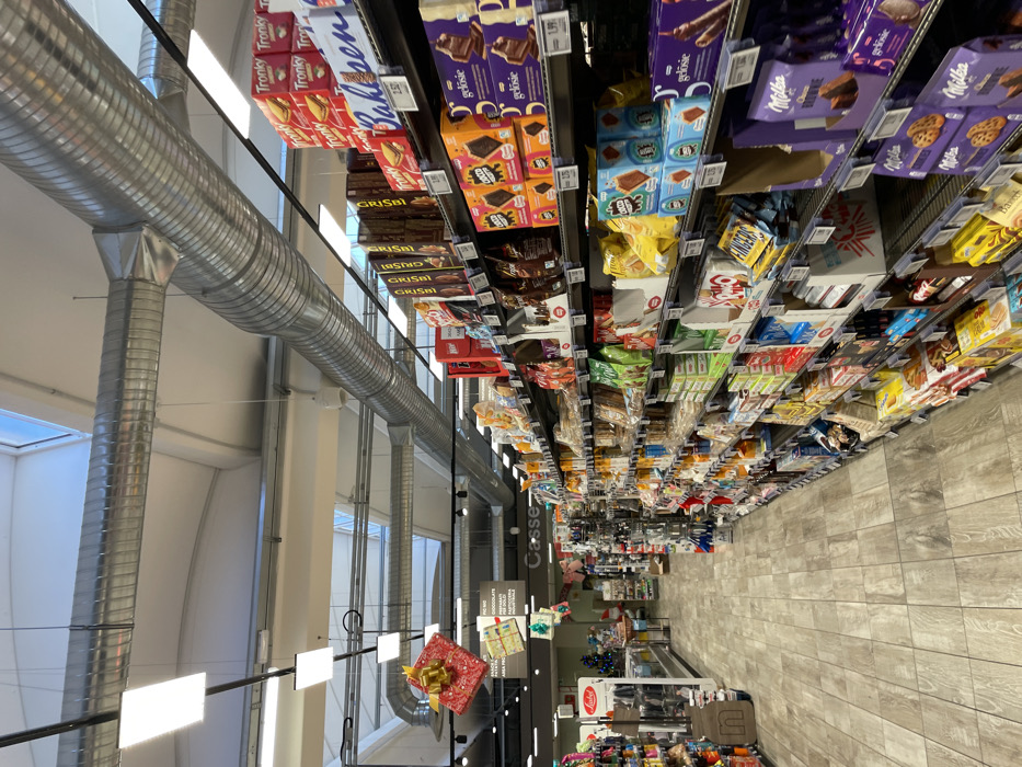 This image depicts an aisle inside a grocery store or supermarket in Castel del Piano, Italy. The scene is bright and well-lit, showing numerous shelves stocked with a variety of packaged food products, primarily chocolates, cookies, and snacks. No people are visible, emphasizing the merchandise and store layout.
The main subjects are the extensive displays of products on metal shelving units. Visible brands include "Tronky," "GRISBÌ," and "Milka," with "BROWNIE" specified for a Milka product. Other discernible brand names are "gelösie" and partially visible "Balisen." Price tags like "1.99" and "2.52" are attached to the shelves.
Notable details include the store's modern industrial aesthetic, characterized by exposed metallic HVAC ductwork running across the high ceiling. Bright illumination comes from both artificial ceiling lights and natural light entering through skylights or large windows on the left side of the frame. The floor is covered with light-colored, patterned tiles. A red, gift-wrapped box with a gold bow hangs from the ceiling, suggesting a festive period or promotional display. Further down the aisle, a small Christmas tree is visible near a sign for "Casse" (Italian for checkouts). Other hanging signs include partial text like "PIC NO CHOCOLATE" and "AREA PRODOTTI," reinforcing the Italian setting. The overall activity is the display of goods in a retail environment.