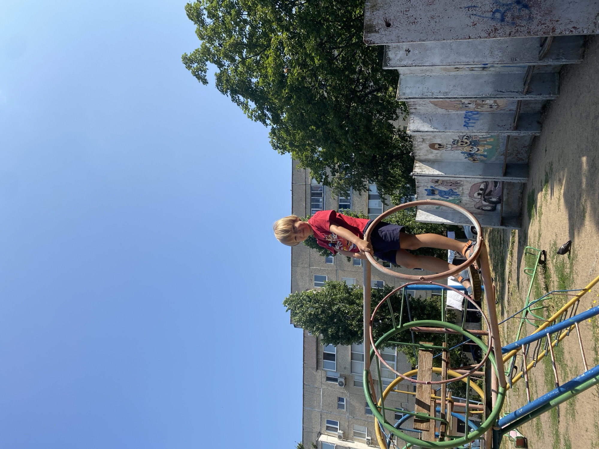 A young blond boy with a red t-shirt and blue shorts is standing on a playground structure in Pivdennoukrainsk, Ukraine. He is holding onto a circular metal bar as he looks at the camera and smiles. The playground is colorful and appears to be made of metal and wood, with various swings and slides. In the background, a grey, multi-story apartment building is visible, along with trees and a clear blue sky. The ground is covered with sand and grass. The overall scene depicts a sunny day in a residential area.