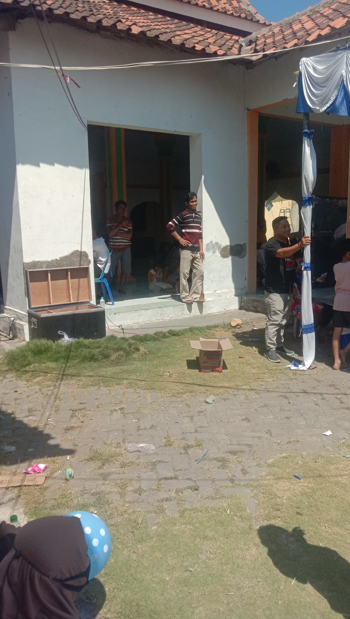 The image depicts an outdoor scene in Karang Anyar, Indonesia, likely in front of a residential building with a tiled roof and white walls. Several individuals are present, with varying levels of activity. In the foreground, a person wearing a brown head covering and a blue polka-dotted object is partially visible. To the right, a person in a black t-shirt and grey pants is standing and adjusting a white fabric draped from a tent structure. Behind them, a child in a pink shirt and dark shorts is also visible.
In the middle ground, a person in a striped t-shirt and beige pants is standing on a raised threshold. Another person in a striped t-shirt and shorts is visible inside a doorway. Further back, several other individuals are seated or standing within the building's entrance. An open wooden box with metal latches sits on the grass to the left, next to a larger black case. A discarded cardboard box with writing on it is placed on the paved area in the center. The ground is a mixture of paved stone and dry grass, with scattered litter including plastic bags and bottles. The overall atmosphere suggests a casual gathering or event.