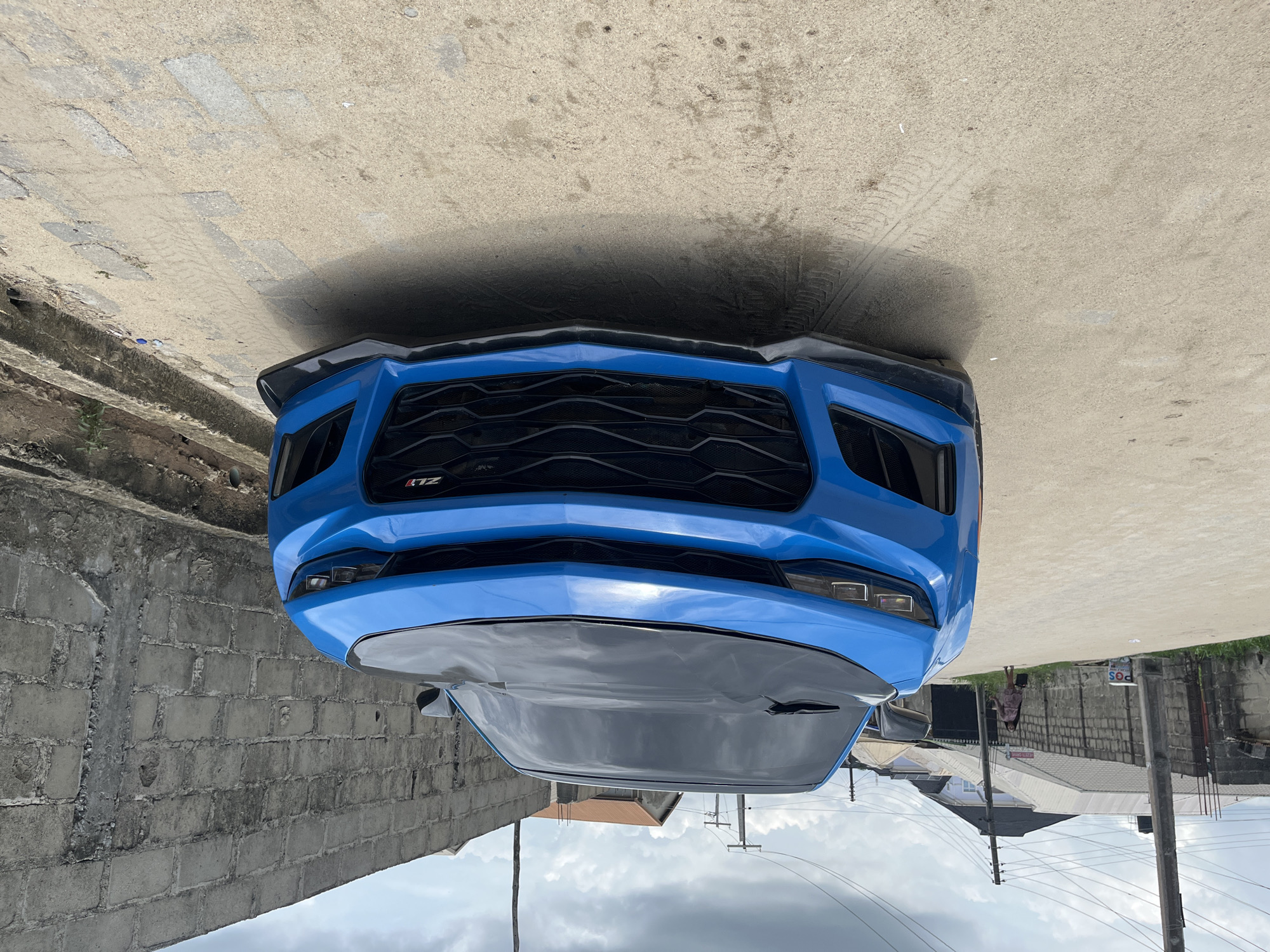 A vibrant blue Chevrolet Camaro ZL1 sports car is prominently featured, seen from a head-on, slightly low-angle perspective. The car is stationary on a concrete surface, with its aggressive front fascia, large black mesh grille, and part of the hood visible. The "ZL1" badge, in red and silver, is identifiable on the lower left side of the grille. The vehicle features black accents on its front splitter and a dark-colored or black-wrapped section on the hood. Its headlights are clearly visible.
To the left of the car, a concrete block wall forms part of the immediate background. To the right, the scene opens to reveal a wider view of the surroundings, which includes more concrete block walls, distant buildings with corrugated metal roofs, and several utility poles carrying overhead wires. The sky appears partly cloudy or overcast, its light reflecting softly off the car's surface.
In the far distance on the right side of the frame, a person, likely a woman, is seen walking away from the car, carrying what appears to be a handbag. A small sign with the letters "POS" is visible near one of the utility poles. The concrete ground in front of the car shows subtle tire marks. The setting indicates a daytime scene in an urban or semi-urban environment, aligning with the provided location context of Iranla, Nigeria.