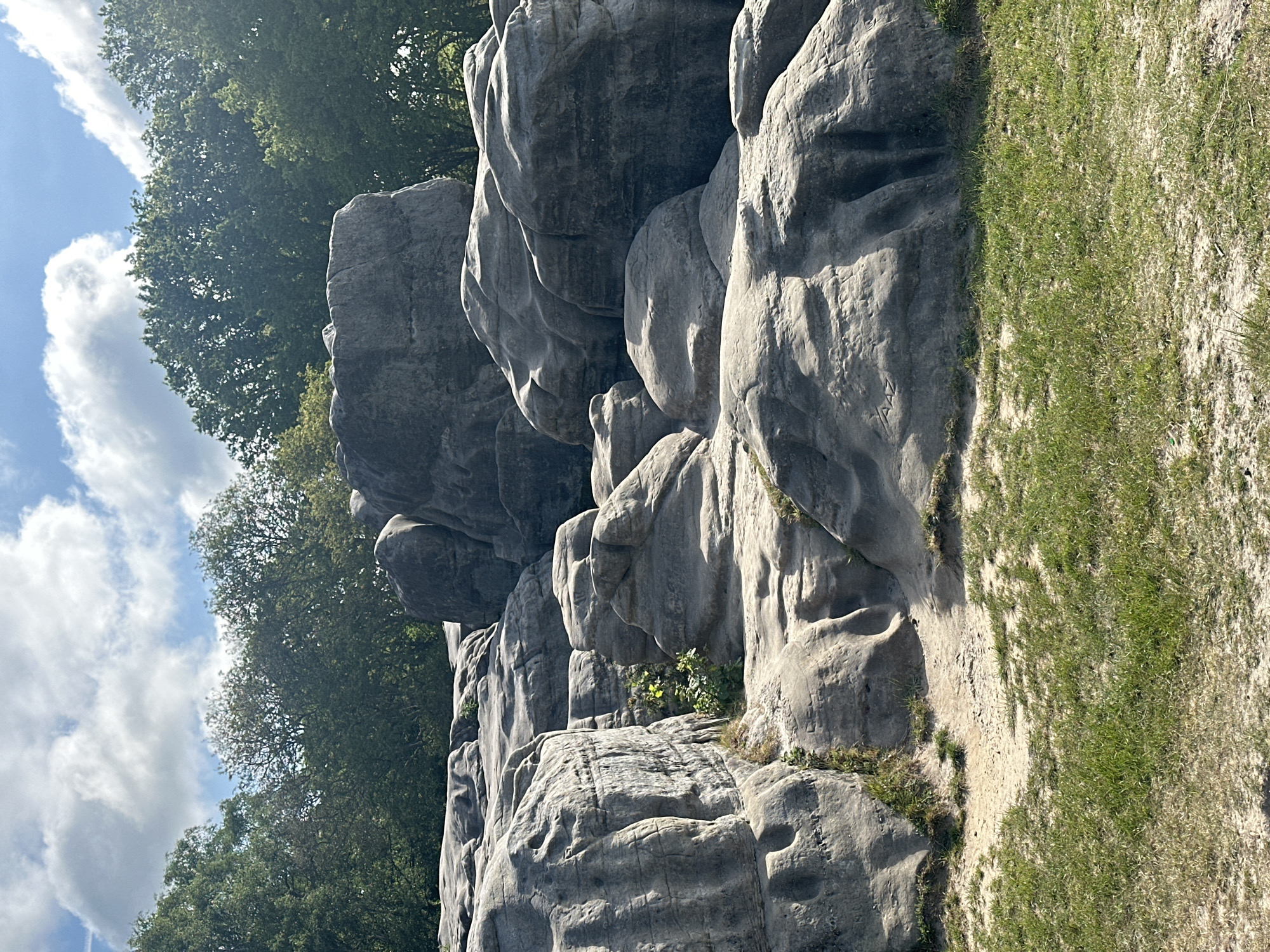 The image depicts a rock formation within the landscape of Tunbridge Wells, United Kingdom. The rocks are light gray, textured, and stacked, exhibiting weathering patterns. Trees with green foliage are visible in the background, reaching towards a sky with scattered white clouds and patches of blue. To the right of the rock formation is a section of grassy terrain and sandy soil.