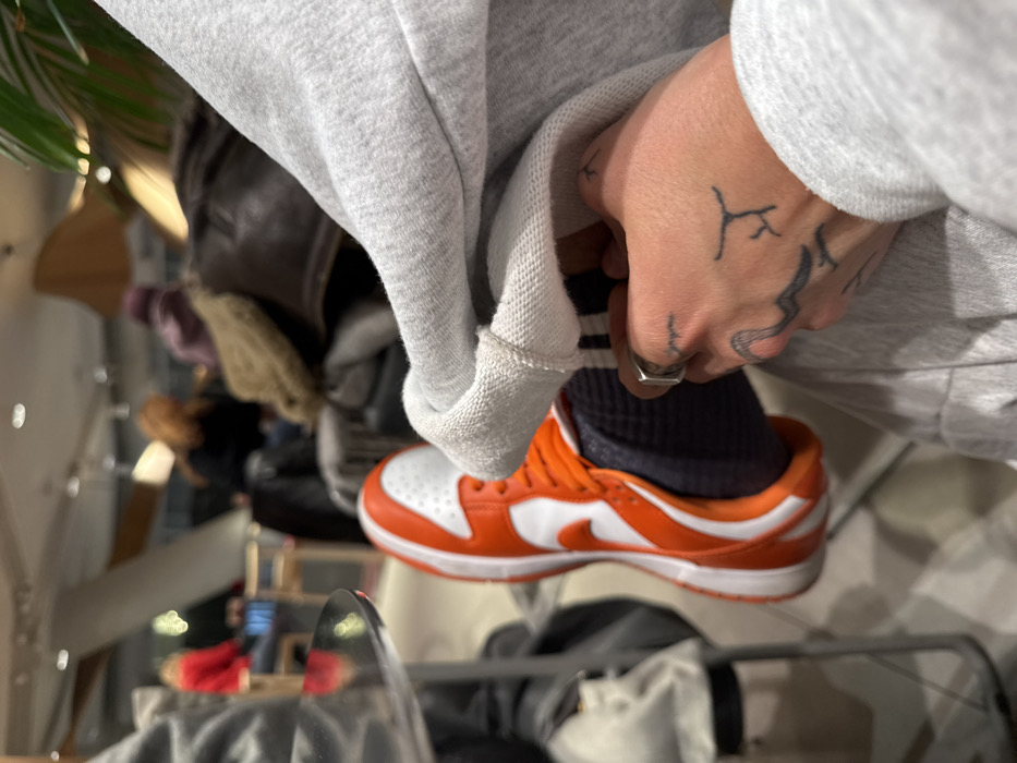 The image features a person wearing orange and white Nike Dunk Low sneakers with dark socks, pulling up the hem of a light gray sweatshirt. The person's left hand is visible, showing several tattoos and a silver ring. The background is blurry, with glimpses of furniture, clothing racks, and other people. The photo is taken at an unusual angle, giving it a slightly disorienting perspective.