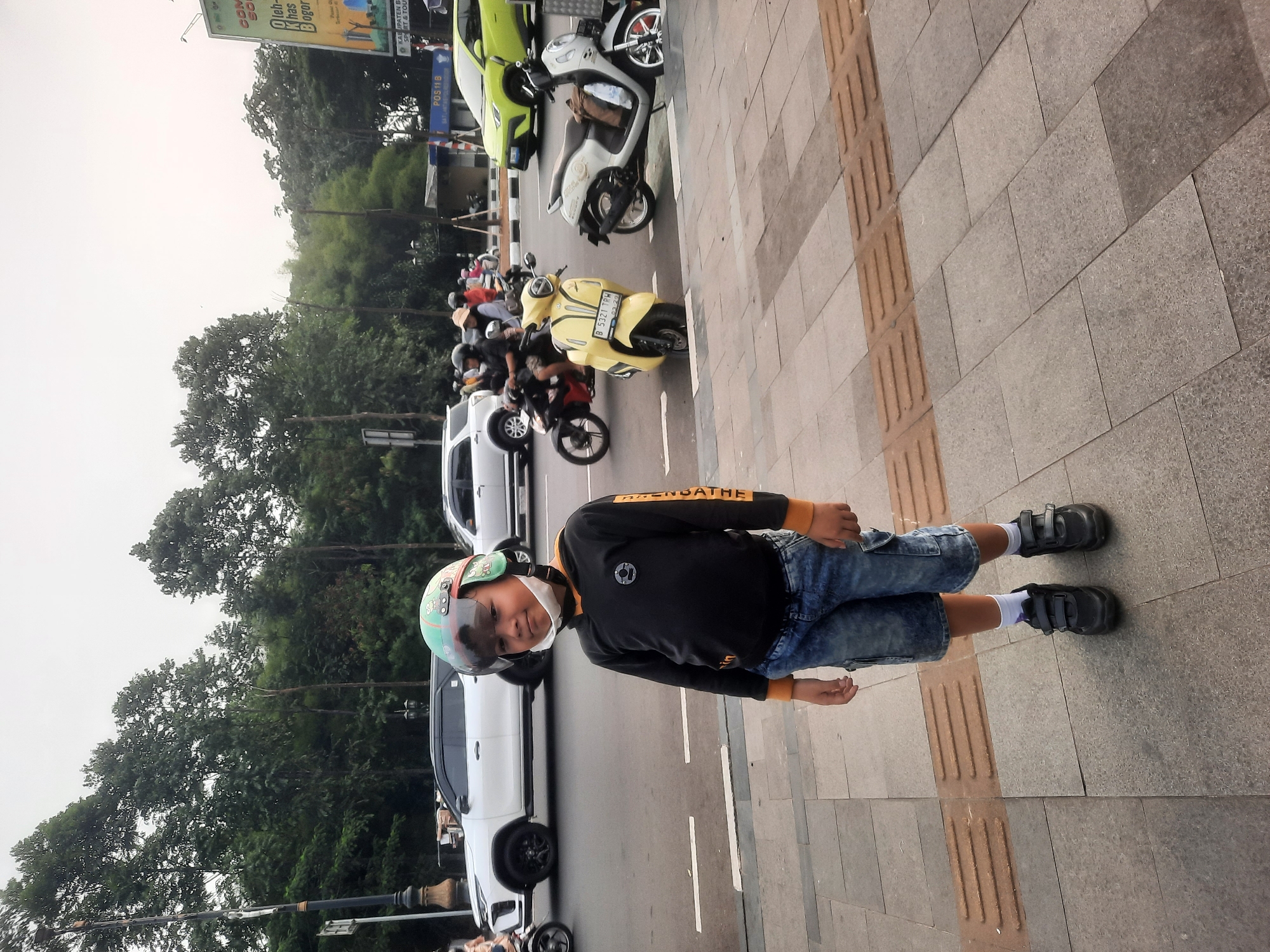 The image depicts a young boy standing on a sidewalk next to a road in Sukaraja, Indonesia. The boy is wearing a black long-sleeved shirt with orange accents on the sleeves, denim shorts, white socks, and black velcro sneakers. He is also wearing a green and white helmet with a face shield. In the background, there are several vehicles on the road, including cars and motorcycles. Trees and foliage line the street, and a billboard is visible in the distance. The sky is overcast. The sidewalk is paved with stone tiles and features tactile paving for visually impaired pedestrians.