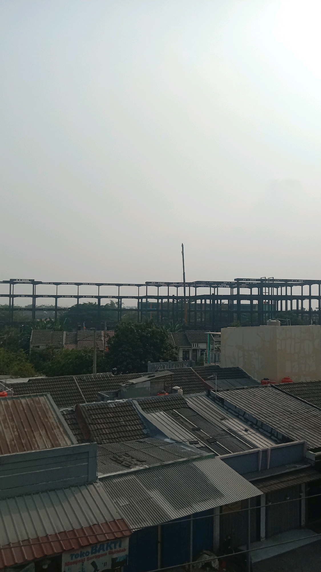 The image shows a suburban landscape in Jalenjaya, Indonesia, under a pale, hazy sky. In the foreground, a collection of residential rooftops are visible, characterized by varying materials including corrugated metal, clay tiles, and some weathered surfaces. Some rooftops feature water tanks. A building with a tan facade stands on the right, partially obscuring the view of some houses. In the middle ground, a densely packed area of homes with red-tiled roofs is interspersed with lush green trees. Beyond this, a massive, unfinished steel structure dominates the background, resembling a large industrial building or stadium under construction, with a tall crane positioned within its frame. The overall impression is one of ongoing development and urban sprawl.