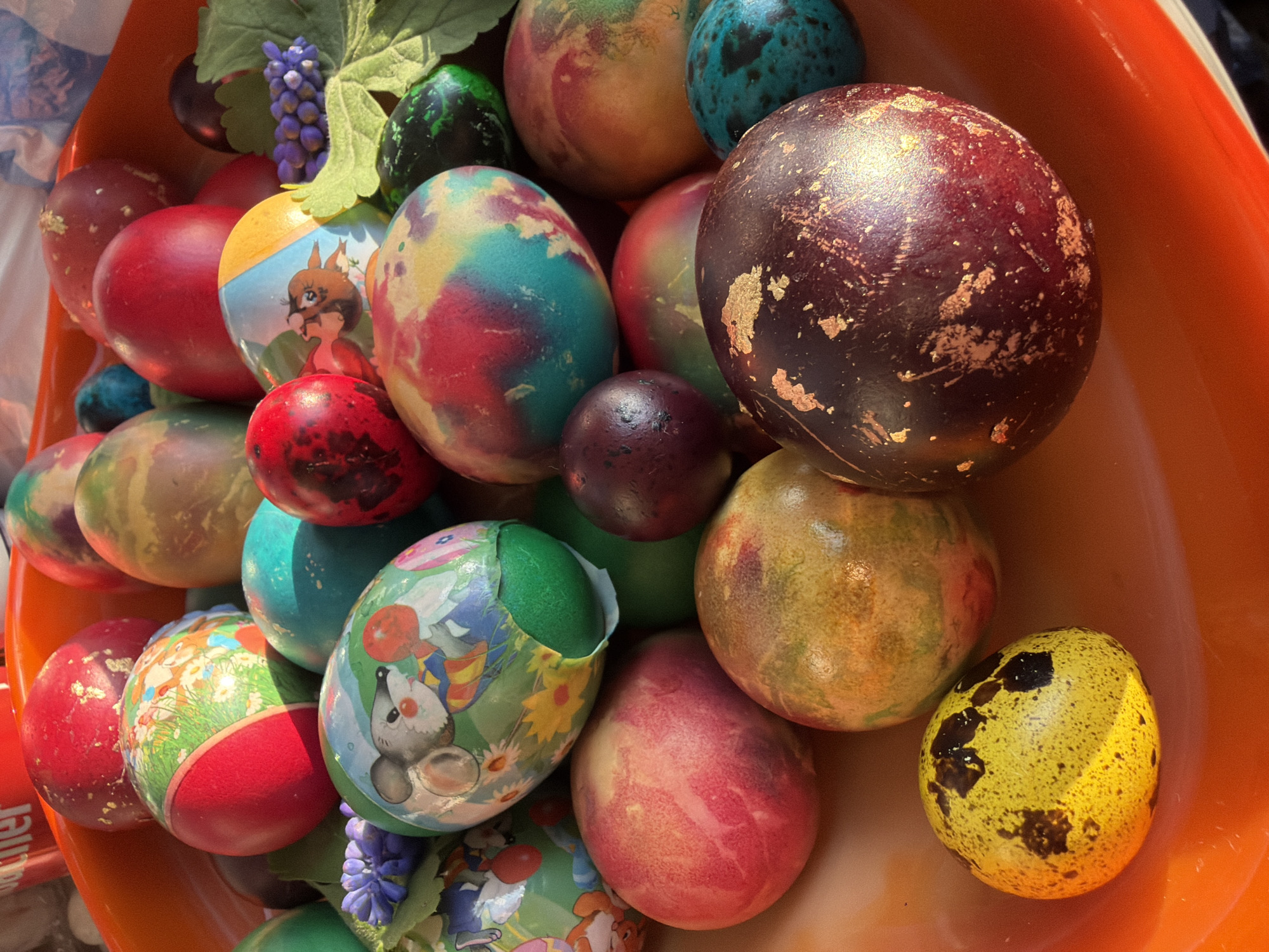 The image shows a collection of Easter eggs in an orange container. The eggs vary in color and decoration, including solid red, yellow with black speckles (resembling a quail egg), tie-dye patterns of red, blue, and yellow, and eggs wrapped in decorative paper with cartoon animal designs. Artificial foliage, including leaves and small purple flower clusters, is also present among the eggs. The container seems to be made of plastic. The lighting is bright and appears to be sunlight. There is no information to confirm the location of the photo in Silistra, Bulgaria.