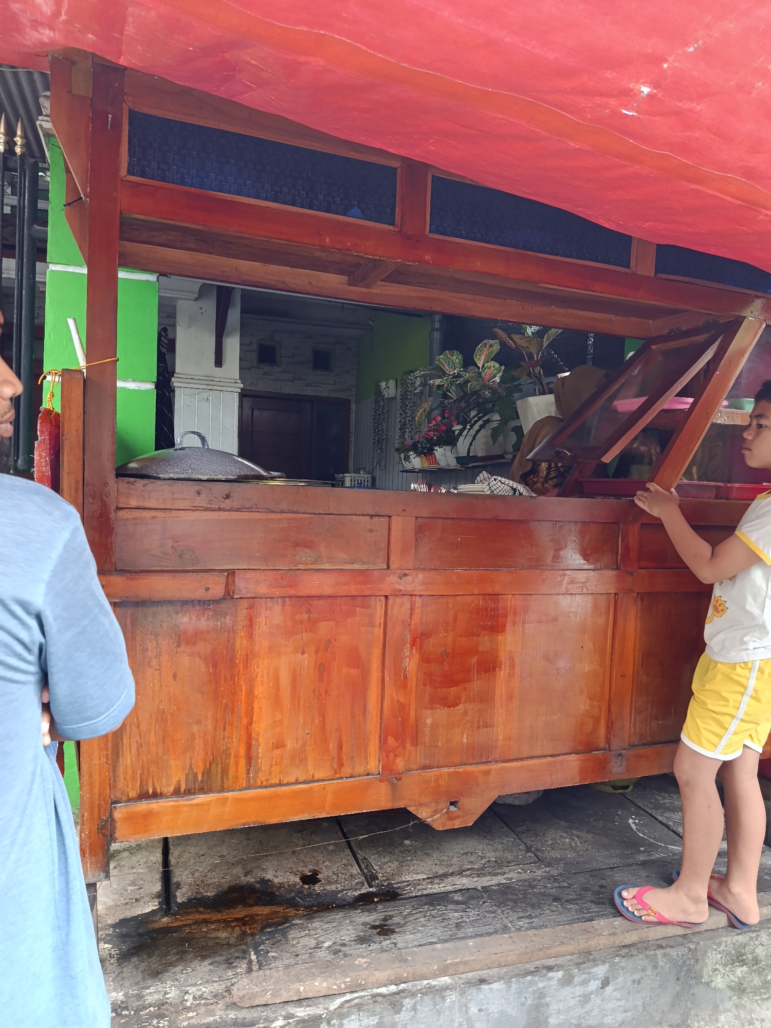 An exterior medium shot captures a wooden food stall in Keboansikep, Indonesia. A red tarp is stretched over the stall, providing shade. The stall is constructed from dark, polished wood, with a large pot with a lid visible on the counter. To the left, a person wearing a light blue t-shirt stands with their back to the camera, partially obscuring the stall. To the right, a child in a white t-shirt with yellow trim and yellow shorts stands facing the stall, looking inside. The child is wearing pink and blue flip-flops. The stall is set up outdoors on a weathered wooden platform, with visible puddles and debris on the ground. In the background, a house with green walls and a patterned window is visible.
