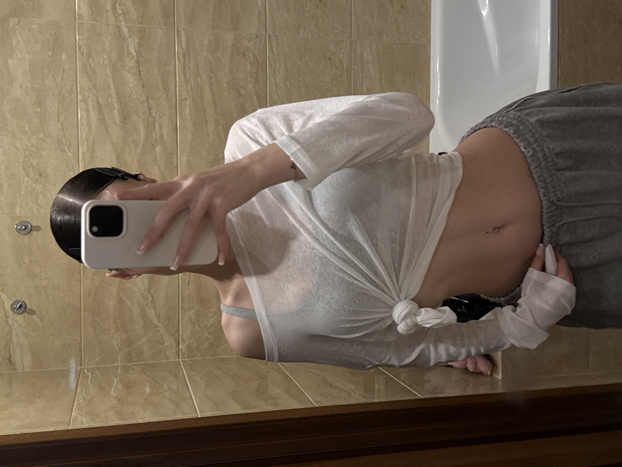 A person is taking a mirror selfie in a bathroom. The person is wearing a white, off-the-shoulder top that is knotted at the waist and grey sweatpants. They have dark hair pulled back and are holding a white smartphone to obscure their face. The bathroom has beige tiled walls and a white bathtub. The scene is set in Khimki, Russia.