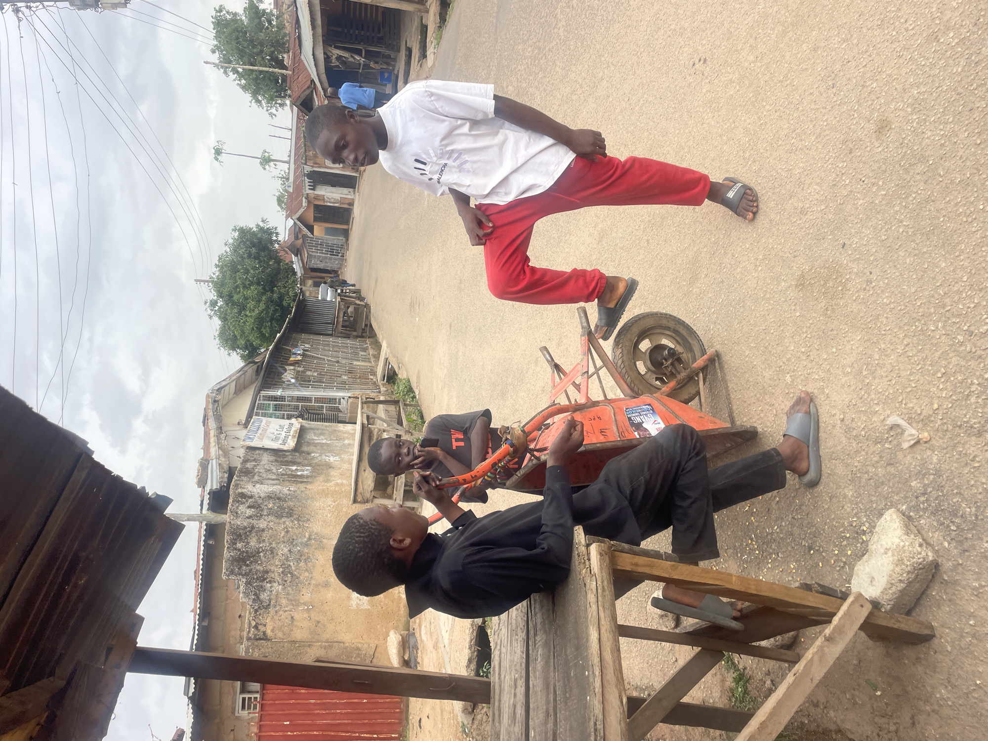 In Jos, Nigeria, an eye-level, outdoor shot shows three boys on a street with buildings on both sides. One boy in a white T-shirt and red pants stands on an orange wheelbarrow, legs spread, looking down. Two boys are closer to the camera; one in a black shirt sits on a wooden structure, and another boy in a black T-shirt stands next to him, both interacting with an object. The street is paved and buildings are varied in construction and color. The sky is overcast.