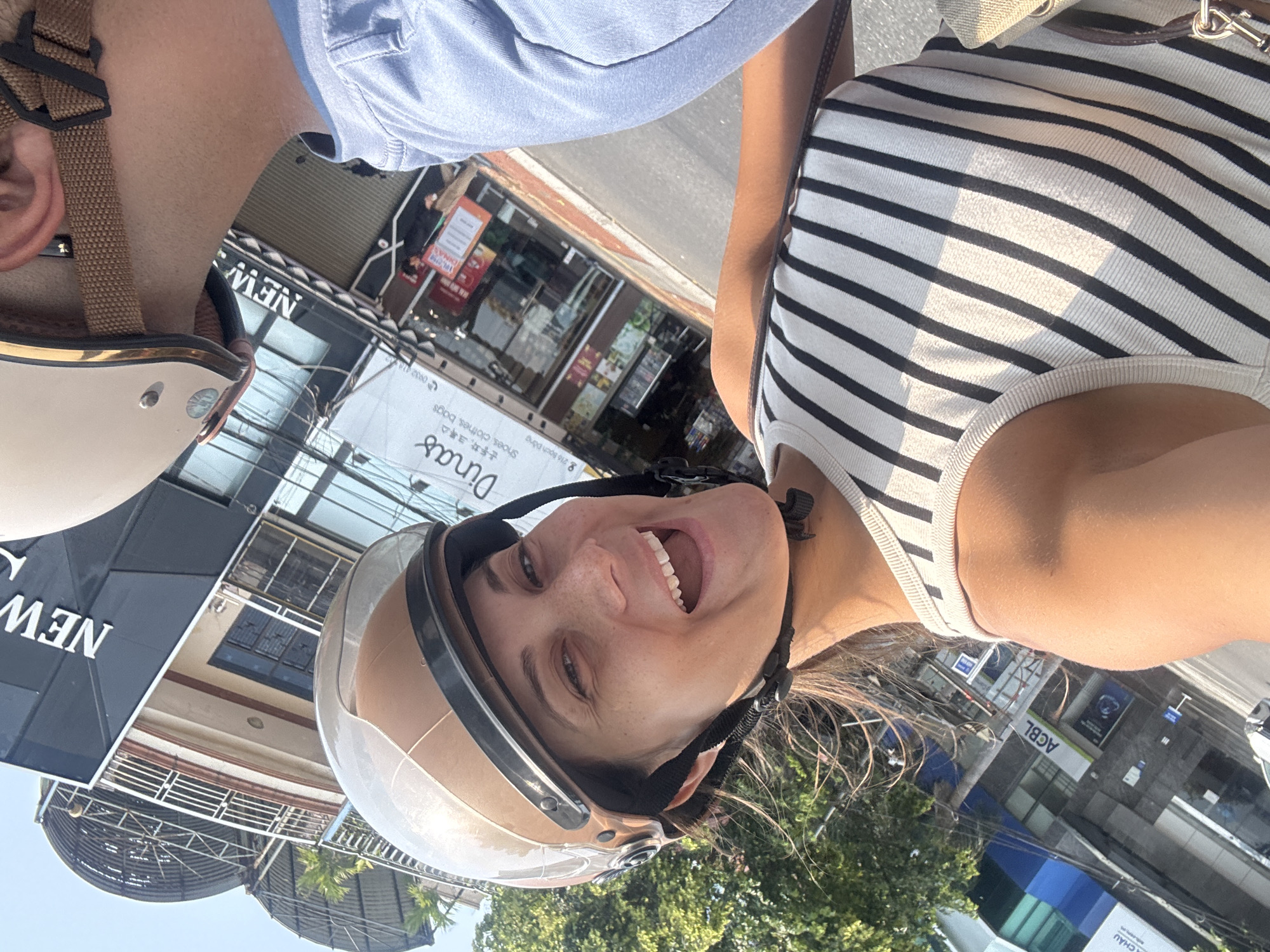 A slightly tilted, eye-level shot captures a woman smiling widely. She is wearing a beige helmet with a clear visor and a striped tank top, seemingly in motion on a motorbike. Adjacent to her is another person wearing a light blue shirt and a similarly colored helmet. The background reveals urban scenery, including storefronts. The image also shows signs in Vietnamese, notably "Dinas" advertising shoes, clothes, and bags. The address "8 216 Bach Dang" is visible, which corresponds to a location in the city of Da Nang, Vietnam. Another sign reads "ACBL." The environment appears to be outdoors, with trees and buildings lining a street.