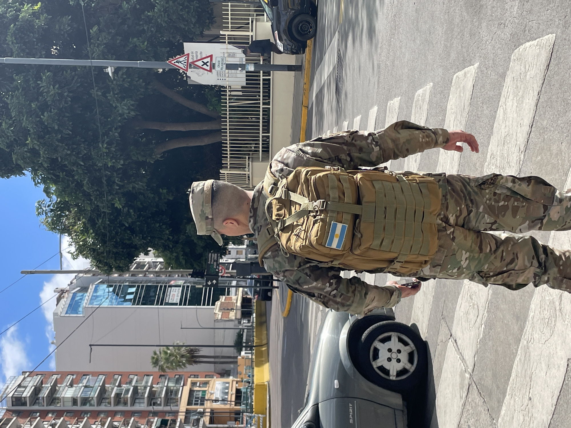 This daylight photograph captures an individual in a military-style camouflage uniform standing on a pedestrian crosswalk in an urban setting. The person, viewed from behind and slightly to the right, wears a matching camouflage cap and a tan tactical backpack. A patch of the Argentinian flag, featuring light blue, white, and light blue stripes with a sun emblem, is prominently displayed on the backpack. Their right hand is visible, holding a small, dark rectangular object, possibly a smartphone or communication device.
The scene unfolds on a paved street with a clearly marked white pedestrian crosswalk. A grey car, with the word "SPIRIT" partially visible on its rear, is positioned to the right of the individual. In the background, multiple tall buildings with reflective glass windows rise, indicative of a city environment. Lush green trees, densely packed, line the left side of the street.
A utility pole on the left features two triangular warning signs: one with a red border and an 'X' symbol, typically signifying a dangerous intersection or level crossing, and another with diverging arrows indicating a complex intersection. Below these, a white rectangular sign has partially visible black text including "CRUCE PELIGROSO" (Dangerous Crossing) and an abbreviation "Avda." followed by numbers "2500 - 2800". The sky above is clear and blue with a few scattered white clouds, suggesting a sunny day. The presence of the Argentinian flag and Spanish text on the signs confirms the location context of Buenos Aires, Argentina. The person appears to be either walking across or waiting at the crosswalk.