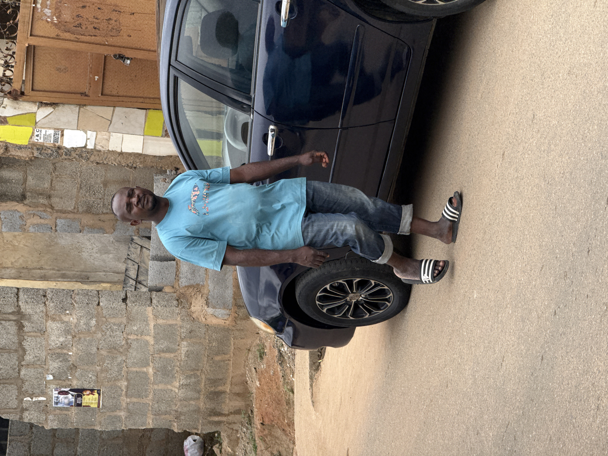 A man is leaning against a dark blue car, his left hand resting on the car door and his right arm extended slightly behind him. He is wearing a light blue t-shirt and blue jeans, with black and white striped slides on his feet. The man has dark skin and a bald head. The car appears to be parked on a dirt or gravel road, with a brick wall and some building structures visible in the background. The overall scene suggests an outdoor setting in a somewhat informal or developing area. The lighting indicates it is daytime. There is no visible text in the image.