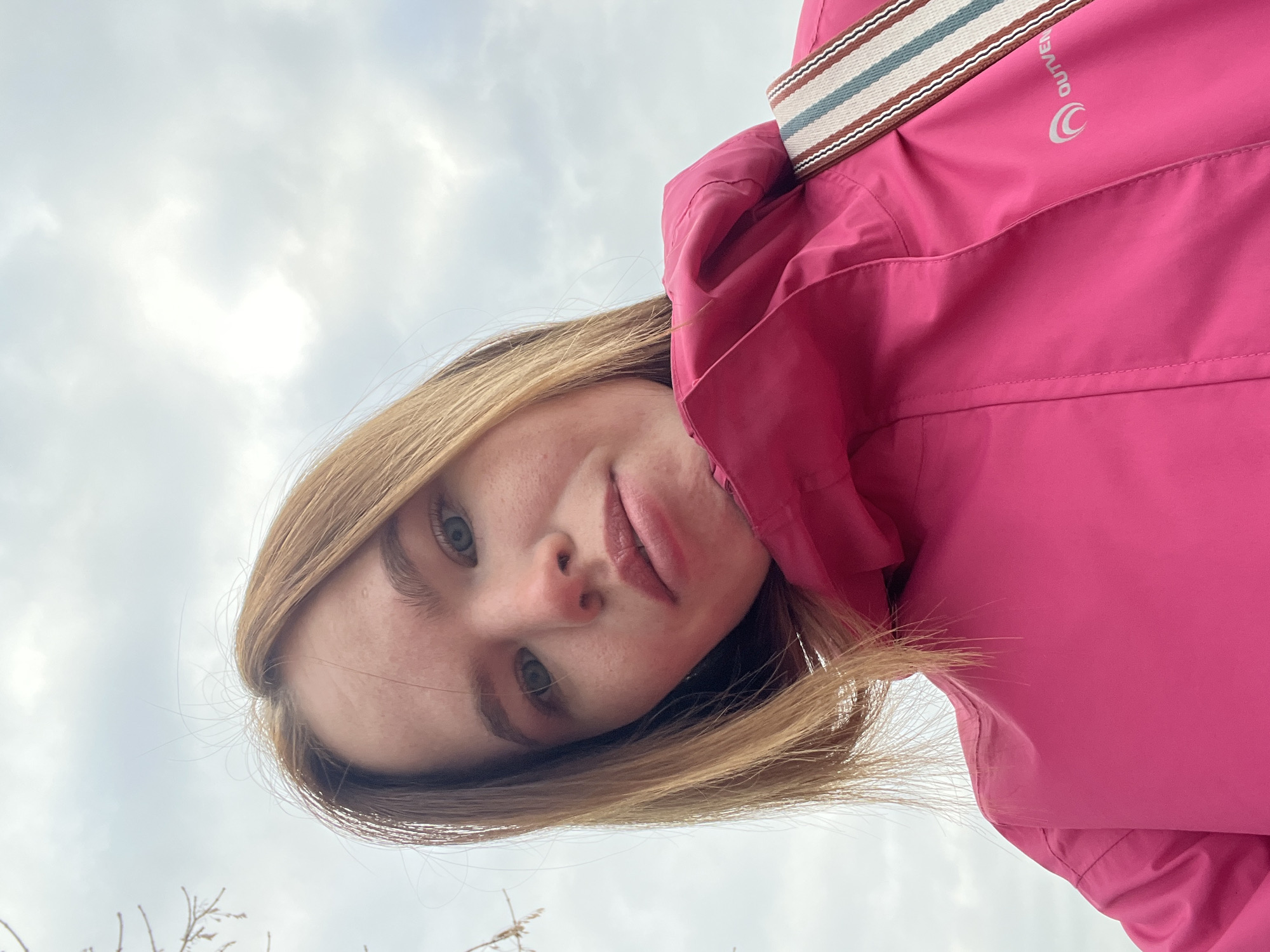 A young woman takes a selfie. She has light brown hair, fair skin and blue eyes. She is wearing a pink jacket and a striped strap across her right shoulder. The background is a cloudy sky and a few branches. The location is unknown and cannot be determined from the image.