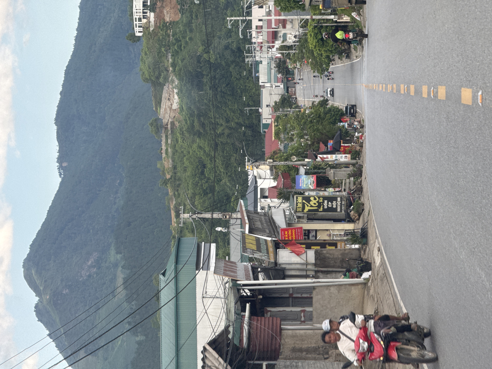 This image depicts a street scene in Tú Lệ, Vietnam, centered on a descending asphalt road. Dense, forested mountains dominate the background under a hazy sky. Power lines are visible traversing the upper left quadrant. Buildings, many featuring corrugated metal roofs in green, red, and gray, line both sides of the road. Vietnamese commercial signage is prominent, including "Tóc" (hair salon) and "Nước Nóng" (hot water). In the foreground on the left, an adult male in a light shirt and dark shorts drives a red motorcycle with a younger passenger wearing a light blue cap. Further down the road, a dark motorcycle with a helmeted rider heads away, and a white car approaches. The road features yellow rectangular reflectors as a center line. A modern white building is visible on a distant, partly cleared hillside to the right. The scene is populated by multiple motorcycles and shows evidence of a semi-urban mountainous settlement.