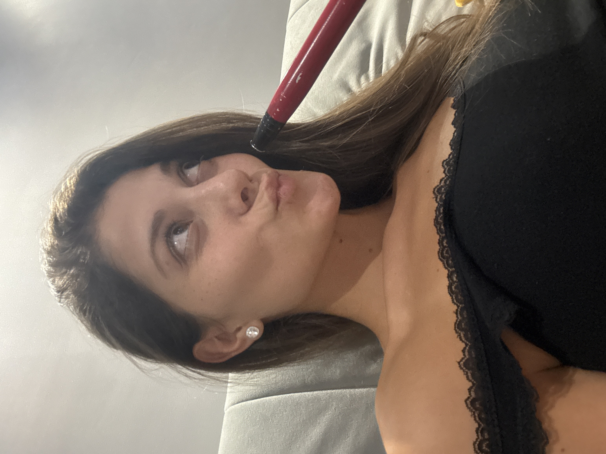 The image shows a close-up of a young woman's face and upper body, taken indoors. She has long, brown hair and is wearing a black off-the-shoulder top with lace trim. She is holding a red object, possibly a hookah hose, to her mouth and looking upwards. Her lips are pursed. She is wearing a diamond stud earring in her left ear. The background appears to be a light-colored couch or wall. There is no discernible location in Burgas, Bulgaria.
