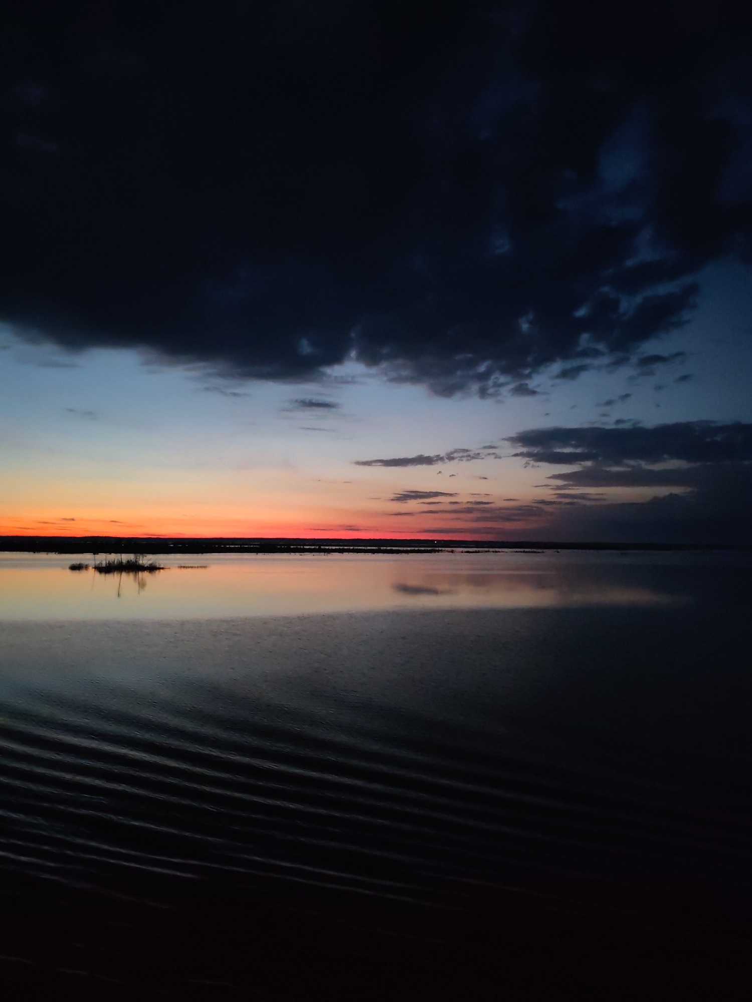 The image depicts a body of water, possibly a lake or river, at twilight or dusk. The sky is dominated by dark, ominous clouds above, transitioning to a gradient of orange, pink, and blue hues closer to the horizon. The setting sun is reflected in the still water. A small, isolated patch of vegetation, possibly an island or cluster of plants, is visible in the distance. Ripples disturb the otherwise smooth surface of the water. The location is unspecified, and the details are too broad to determine if it is in Bereg, Russia. There is no evidence of people or specific objects that can provide a forensic location.
