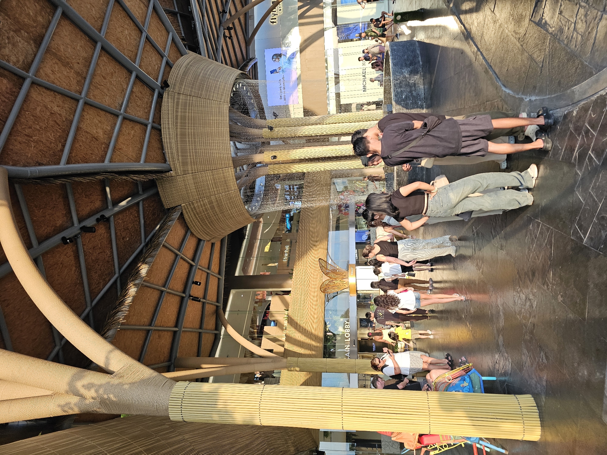 The image captures a sunny outdoor scene at a modern shopping mall or complex in Denpasar, Indonesia. In the foreground, a group of people are seen walking and standing. The architecture features large, bamboo-like columns and a woven textured ceiling structure, with a fountain element cascading water. Various shops are visible in the background, including "MICHAEL K" and "FLYING FISH COFFEE". The ground is paved with dark tiles with a reflective quality. Sunlight casts strong shadows, indicating it is likely late afternoon. The environment appears to be a public gathering space, possibly a central atrium or plaza within a commercial development.