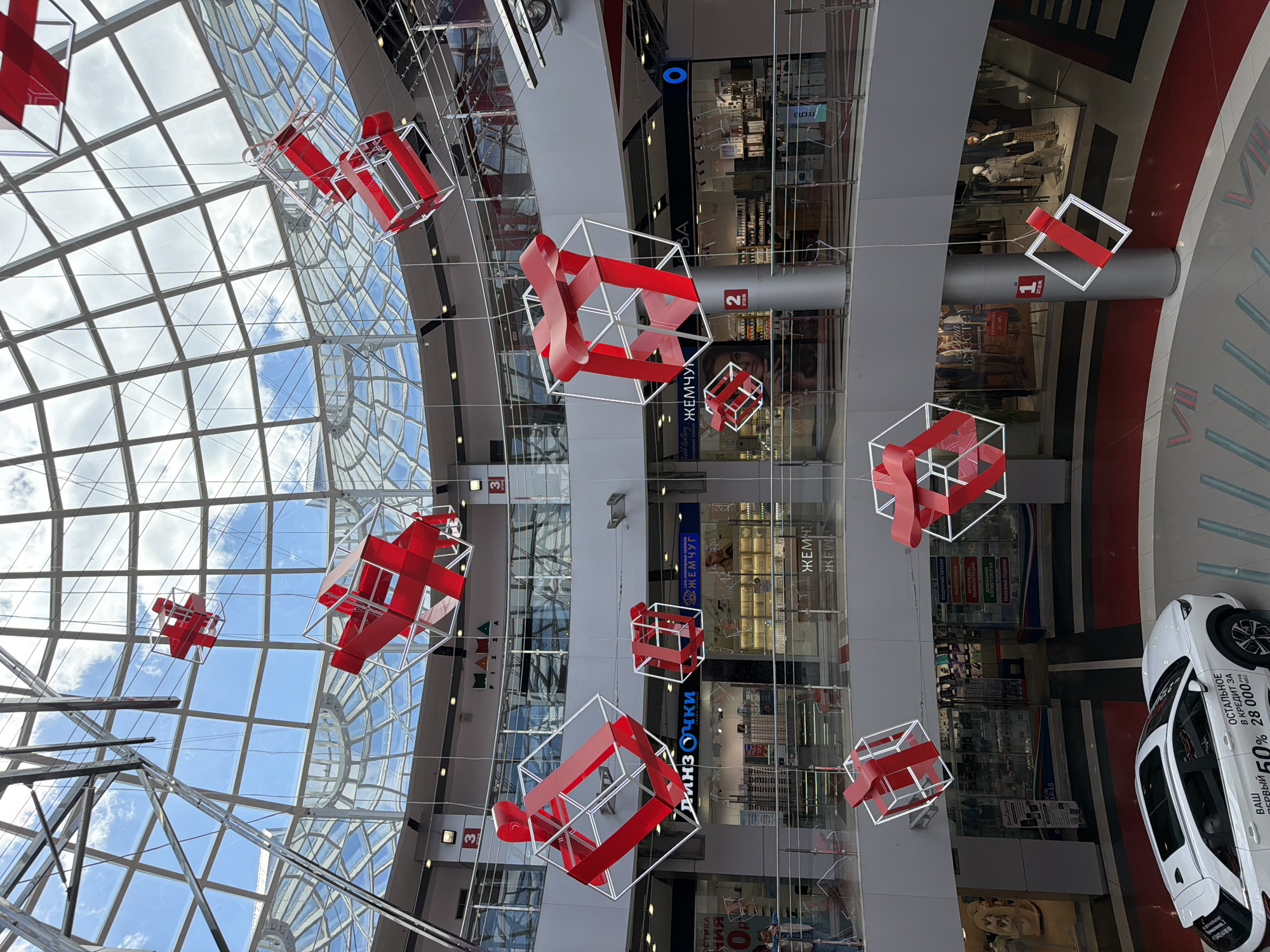 Here's a detailed description of the image:
The image appears to be taken inside a shopping mall, potentially in Izhevsk, Russia. It's a vertically oriented shot looking upwards, emphasizing the mall's interior architecture and decorations.
**Subjects and Objects:**
* **Decorations:** Numerous decorative cube-shaped objects hang from the ceiling. These are constructed with white frames and red ribbon details resembling gift boxes.
* **Mall Structure:** The ceiling consists of a grid-like glass structure allowing natural light to enter. Several floors of the mall are visible with storefronts and signage.
* **Storefronts:** Store signs are visible in Russian, and some storefronts display clothing and other merchandise. Signs indicate locations like "ЖЕМЧУГ" (likely a jewelry store, "Pearl" in English) and "Очки" (Eyeglasses).
* **Car:** A white car is partially visible in the lower right corner. It features promotional graphics in Russian, highlighting a credit offer and a percentage discount (50%).
* **People:** Though no people are clearly visible, mannequins can be seen through the storefront windows.
**Environment:**
* The interior appears modern, clean, and well-lit, thanks to the glass ceiling.
* The overall setting indicates a commercial shopping environment.
**Specific Details Potentially Relevant to Location (Izhevsk):**
* **Language:** The Russian signage strongly suggests a Russian-speaking location.
* **Store Names:** Knowing the specific store names ("ЖЕМЧУГ", etc.) might help pinpoint the specific mall if they are common in Izhevsk.
* **Car Advertisement:** The specific car model and the details of the promotional credit offer, along with the dealership mentioned, could potentially be used to determine its location.