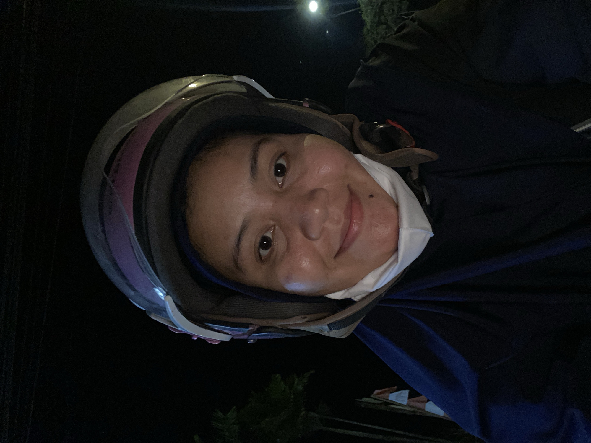 A head-and-shoulders selfie taken at night shows a person wearing a dark blue hijab and a dark blue jacket, with a white face mask hanging below their chin. They are also wearing a gray and pink helmet with a clear visor. The person is looking directly at the camera and smiling. The background is dark, with some blurry green foliage and a distant light pole visible. The scene appears to be outdoors, possibly on a street or roadside, given the presence of foliage and the light pole.