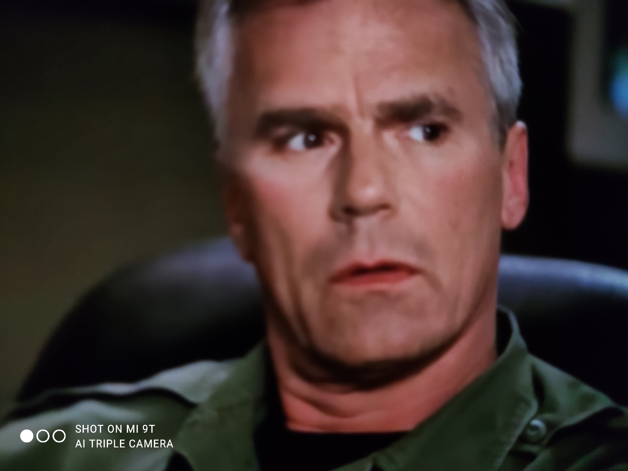 The image is a close-up shot of a man with short, graying hair and prominent eyebrows. He is looking slightly off to his right with an expression of surprise or concern. His mouth is slightly open. He is wearing a dark green, collared shirt over a black undershirt. The background is dark and out of focus, with some faint lights visible on the right side. There is a watermark in the bottom left corner that reads "SHOT ON MI 9T AI TRIPLE CAMERA". The image does not contain any discernible elements that would indicate a location in Khazar Raion, Azerbaijan.