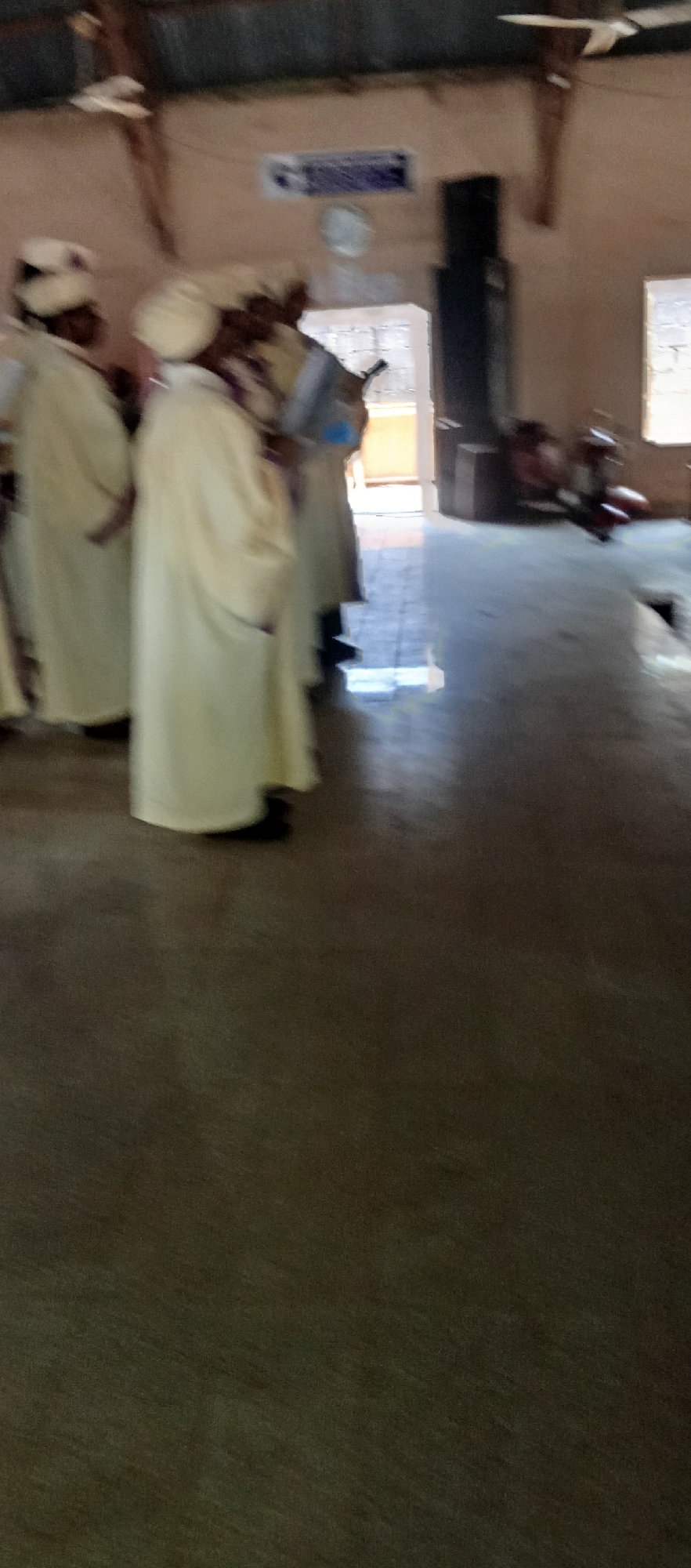The image shows a group of men in white robes and turbans standing inside a building, possibly a church or religious institution, in Akwanga, Nigeria. The building appears to be a simple structure with a metal roof and concrete floor. There is an open doorway in the background, with light streaming in. A loudspeaker, part of a sound system, is visible near the door. A ceiling fan can be seen attached to the roof.