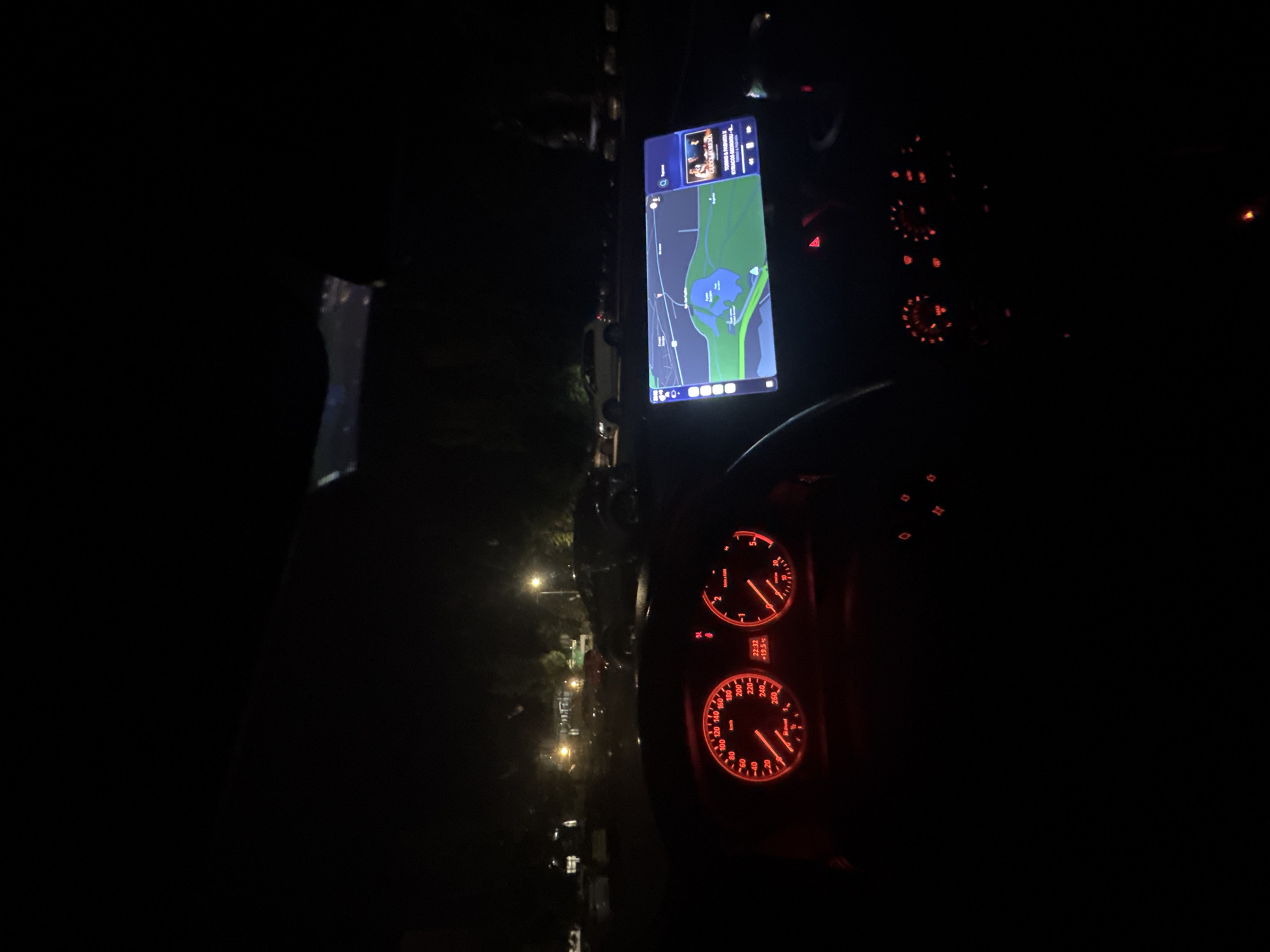 The image shows the interior of a car at night. The dashboard is visible with glowing red gauges, including a speedometer and tachometer. A large tablet-like device is mounted on the dashboard displaying a map with a body of water, possibly a lake, and route guidance. The map appears to indicate a location near Stara Zagora, Bulgaria, as a similar body of water is located there. The exterior shows streetlights and glimpses of the road and parked vehicles. The overall impression is of driving through a dark street in a somewhat urban environment.