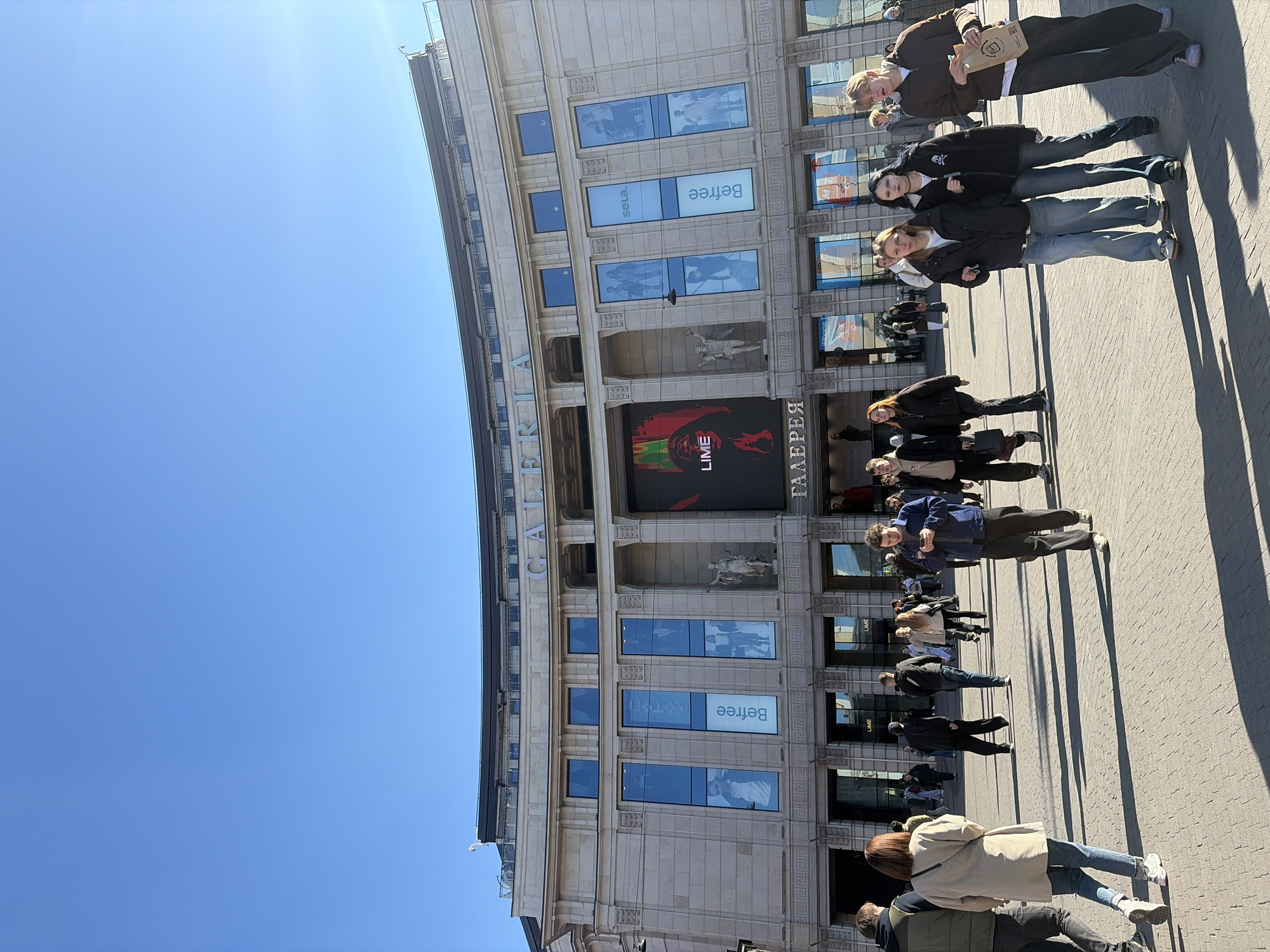 The image shows a street scene in Saint Petersburg, Russia, in front of the Galeria shopping mall. The building is a large, multi-story structure with neoclassical architectural details, including columns, statues, and ornate window frames. Signage for the mall is visible, including window displays with brands like "Sela" and "Befree" and a large LED screen advertising "Lime."
A crowd of people is walking along the sidewalk in front of the mall. The people are wearing casual clothing and appear to be shopping or sightseeing. Shadows suggest it is a sunny day.
