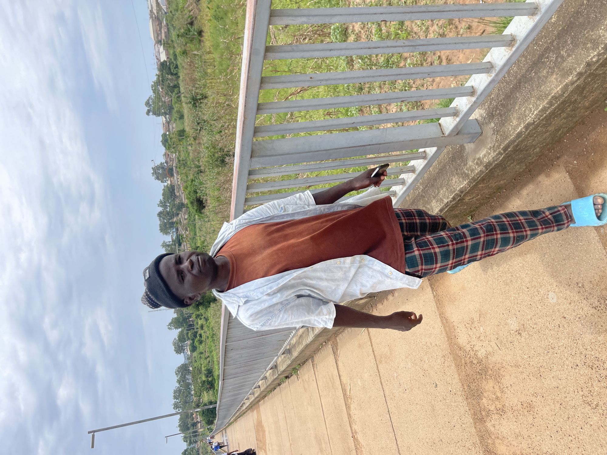 A young Black male wearing a black beanie, a rust-colored t-shirt, an unbuttoned white shirt, and plaid pants walks along a concrete bridge in Rafin Par, Nigeria. The bridge has a grey metal railing. A rural landscape of green vegetation and scattered buildings is visible in the background. The sky is overcast with grey clouds. The man is looking slightly to his right and holding a dark-colored smartphone in his right hand. His left hand is slightly extended with his fingers spread. He is wearing light blue slide sandals. Several other people are walking on the bridge in the distance to the left.