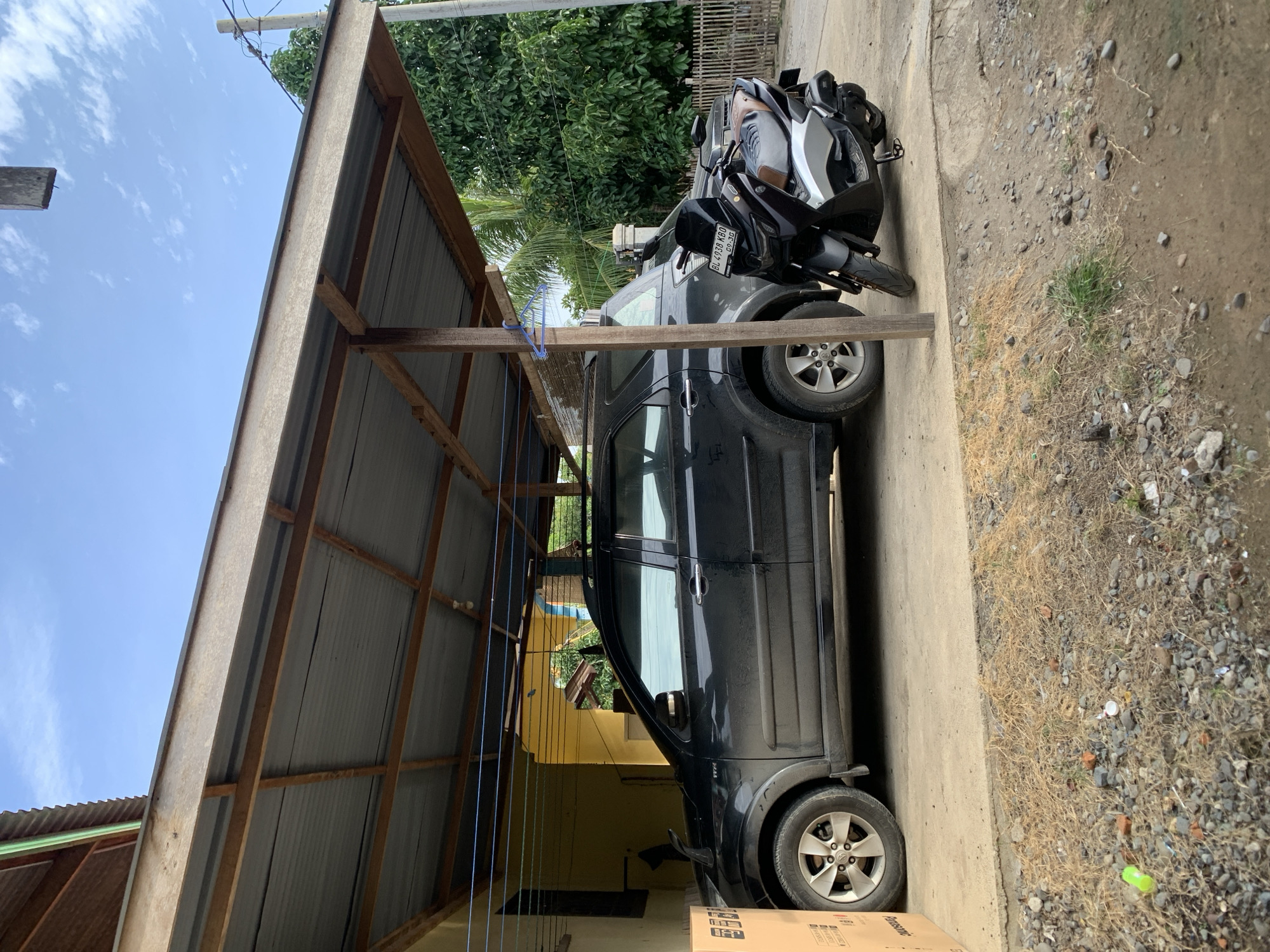 In Meunasahdrang, Indonesia, a black SUV is parked under a simple wooden carport with a corrugated metal roof. A black motorcycle is parked to the right of the SUV. The ground is a mixture of concrete and gravel. In the background, there is a yellow building, lush green trees, and a wooden fence. The sky is blue with scattered clouds.