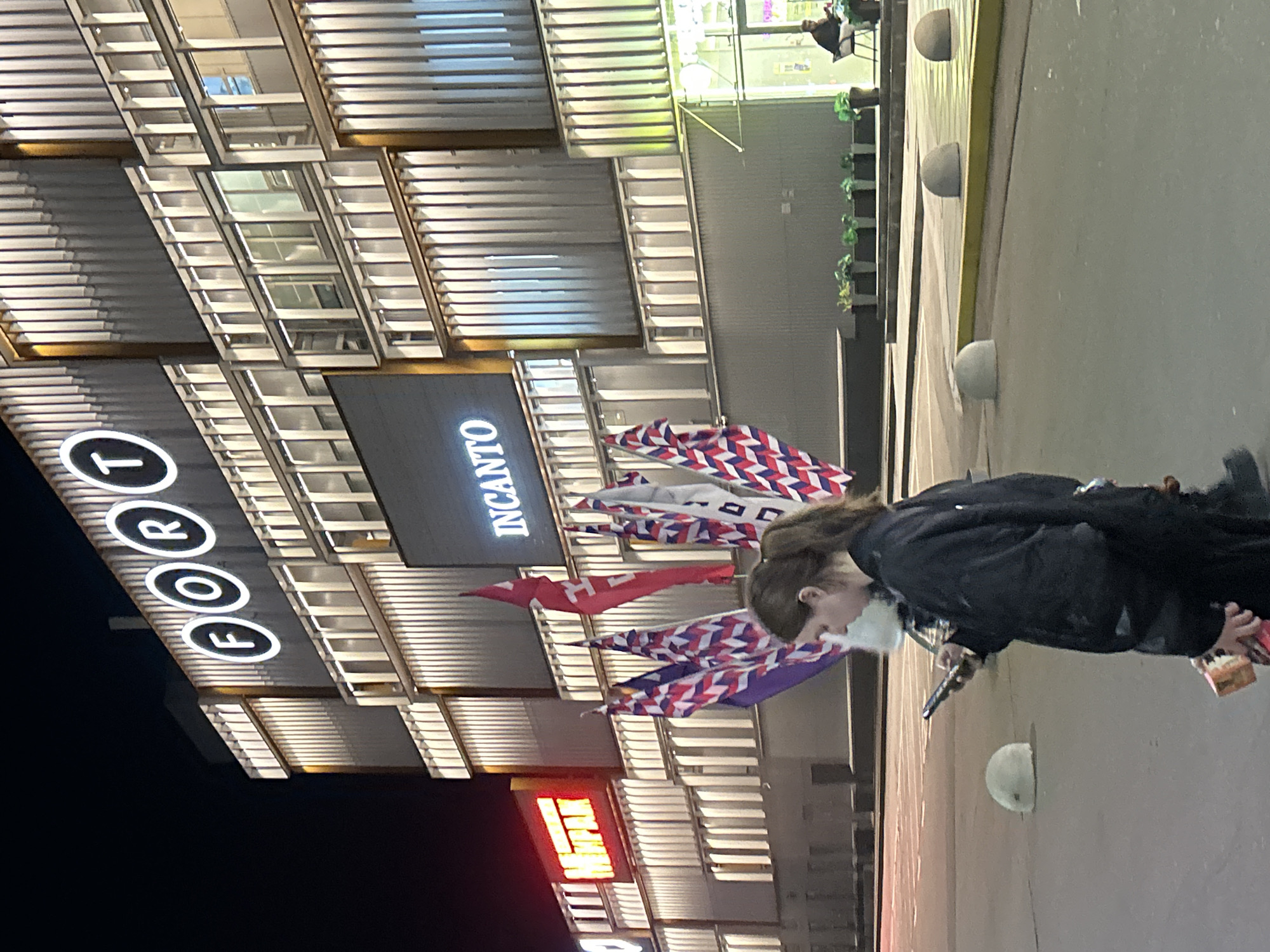 The image shows the "FORT" shopping center and "INCANTО" store in Moscow, Russia, at night. The building has a modern design with illuminated vertical elements and a large screen displaying "INCANTО". Several flags with geometric patterns and a red flag are displayed in front of the building. A young woman in a black jacket is walking and looking at her phone. She is holding a small box and a red item in her hands. In the foreground, there are concrete bollards. In the background, there is a person sitting at a table outside a store entrance.