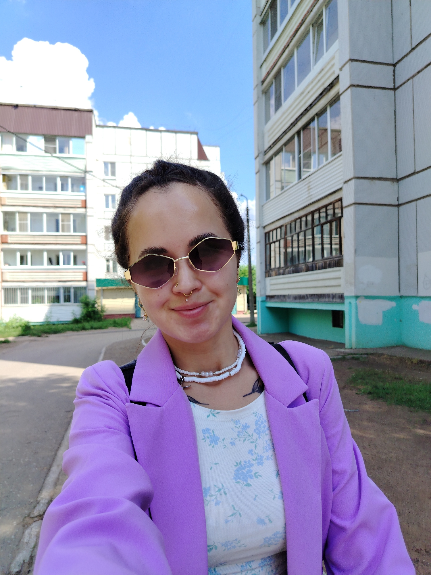 A woman with dark hair, sunglasses, and a nose ring takes a selfie outdoors. She is wearing a purple blazer over a floral top and a white beaded necklace. The background shows two apartment buildings, one on the left with brown and white siding, and another on the right which is taller and has light grey siding with teal accents. The area appears to be a residential street in Chaikovsky, Russia. The sky is blue with some white clouds.