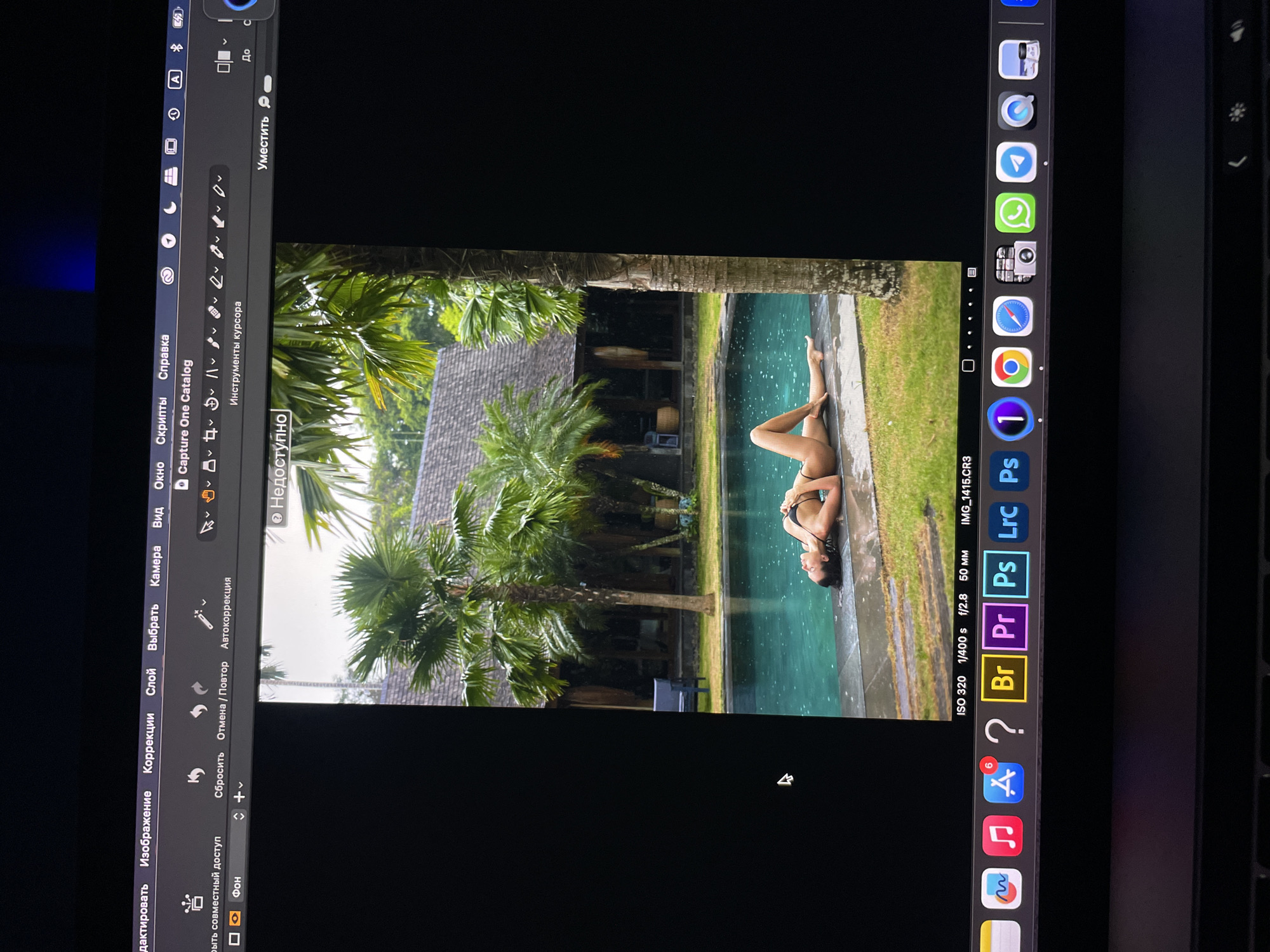 The image shows a laptop screen with a photo editing program open, displaying a photograph of a woman lying by a pool in a tropical setting. The woman is wearing a bikini. The pool is surrounded by green grass and palm trees. In the background, there is a building with a thatched roof. The photo editing program is Capture One, and the icons on the task bar show other editing applications such as Lightroom, Photoshop, and Bridge. The OS is MacOS. There is no indication of Kazan, Russia, in the image.