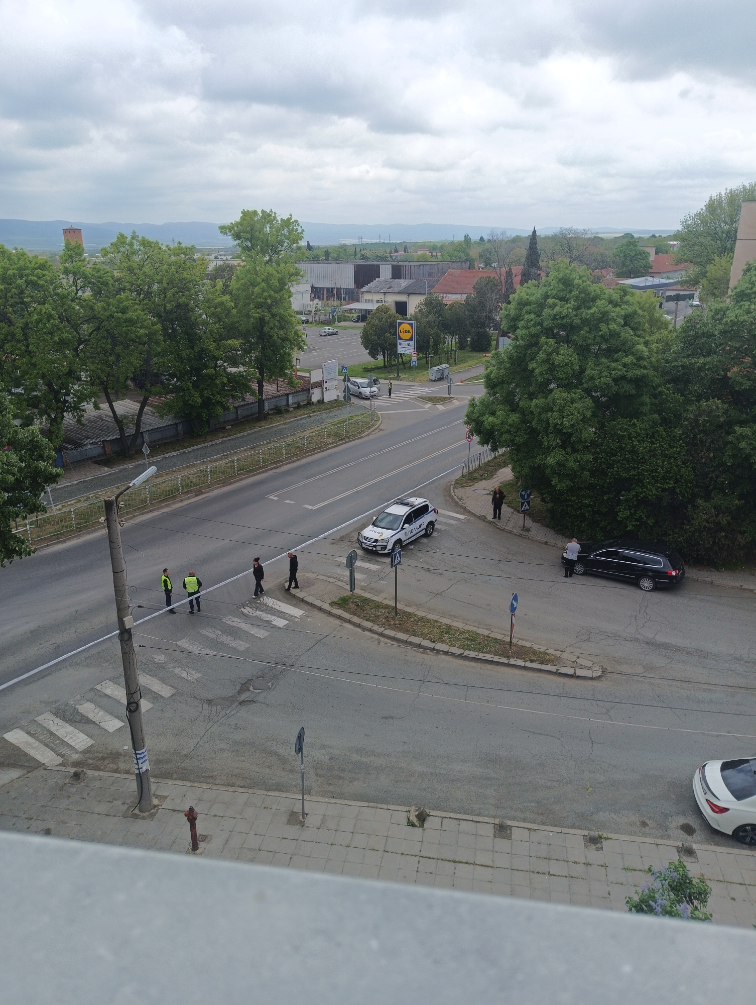 The image shows a street scene in Karnobat, Bulgaria, observed from an elevated viewpoint. Two police vehicles are present, one white and one black, each attended by two individuals. One set of people appears to be inspecting the car. The police officers, clad in high-visibility yellow jackets, are interacting with two pedestrians on a zebra crossing. In the background, a Lidl supermarket is visible.